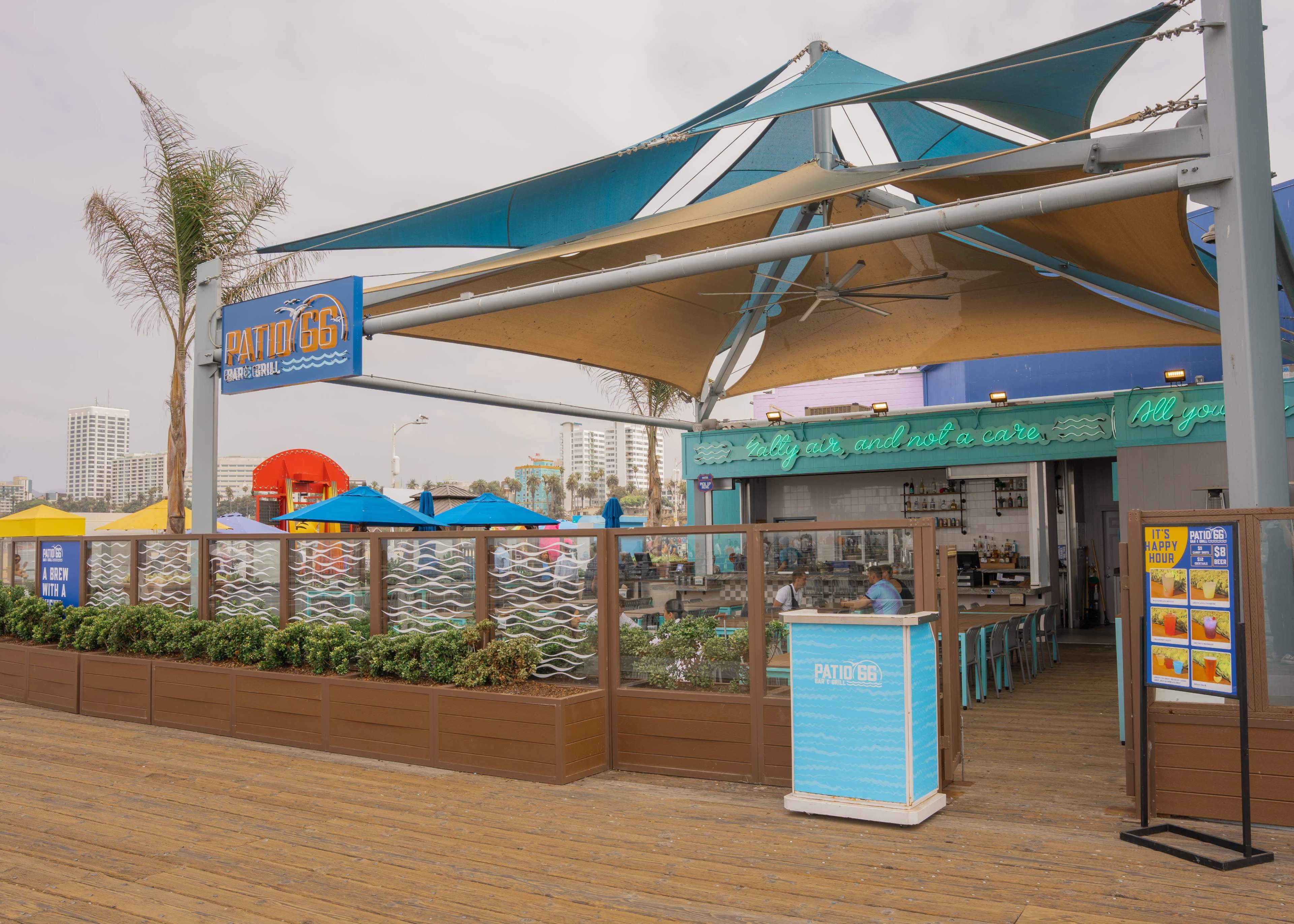 A beachfront restaurant with a vibrant canopy and outdoor seating is shown, surrounded by colorful umbrellas and palm trees.