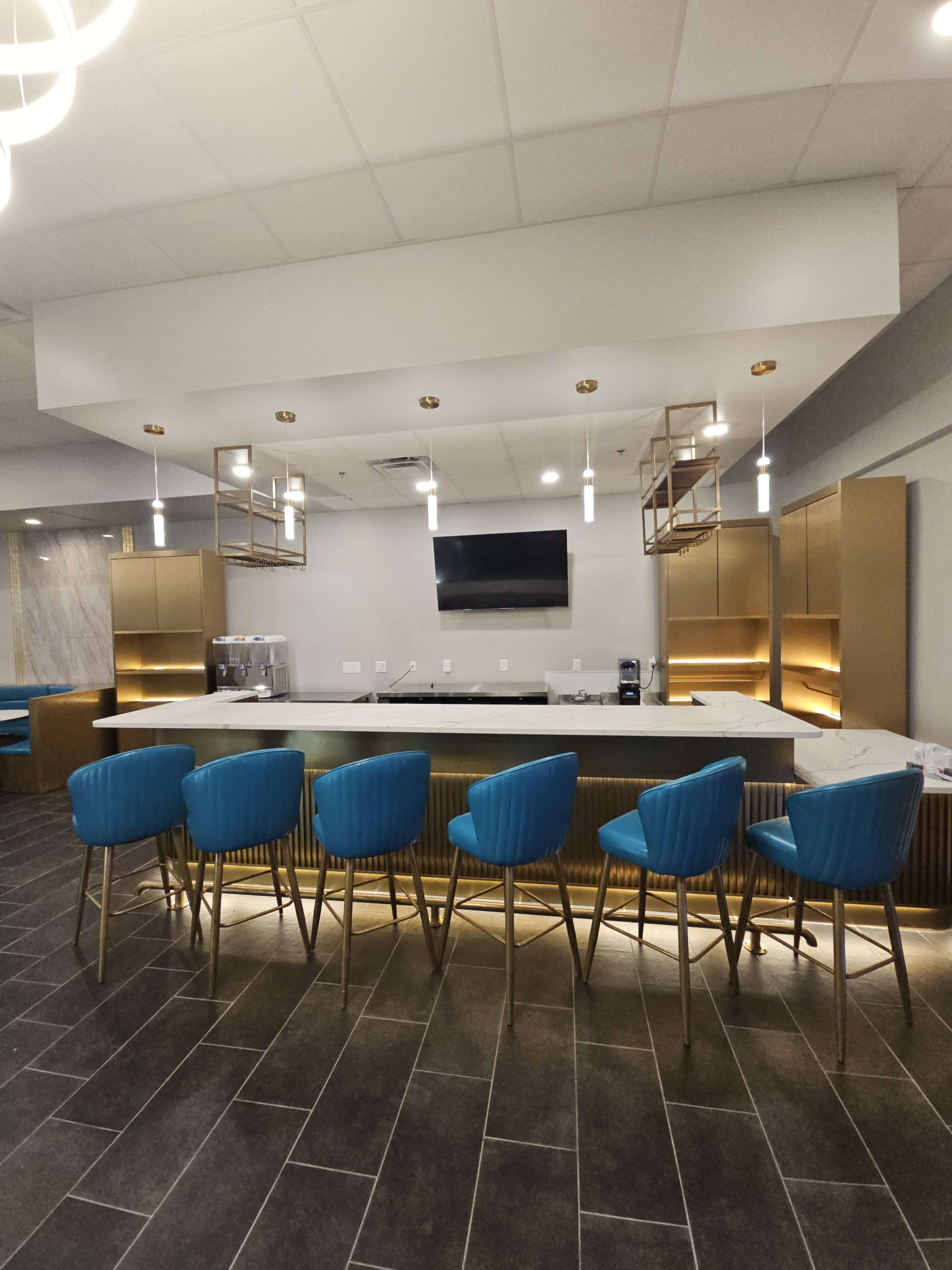 The image shows a modern bar area with blue stools arranged in front of a sleek counter, accented by gold cabinetry and lighting, and a television mounted on the wall.