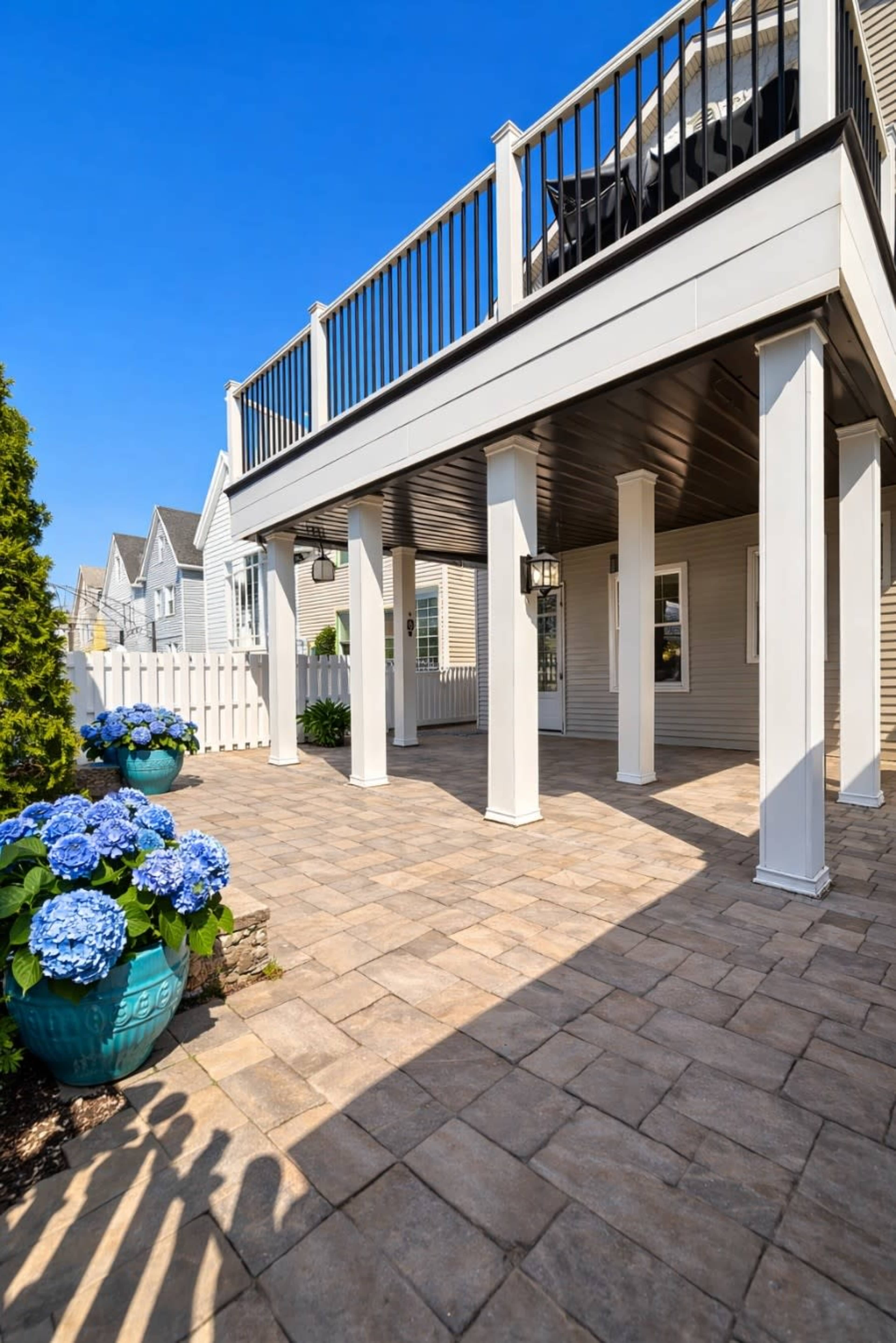 The image shows a patio area with a stone floor, white columns, and a potted hydrangea plant, all under a clear blue sky.