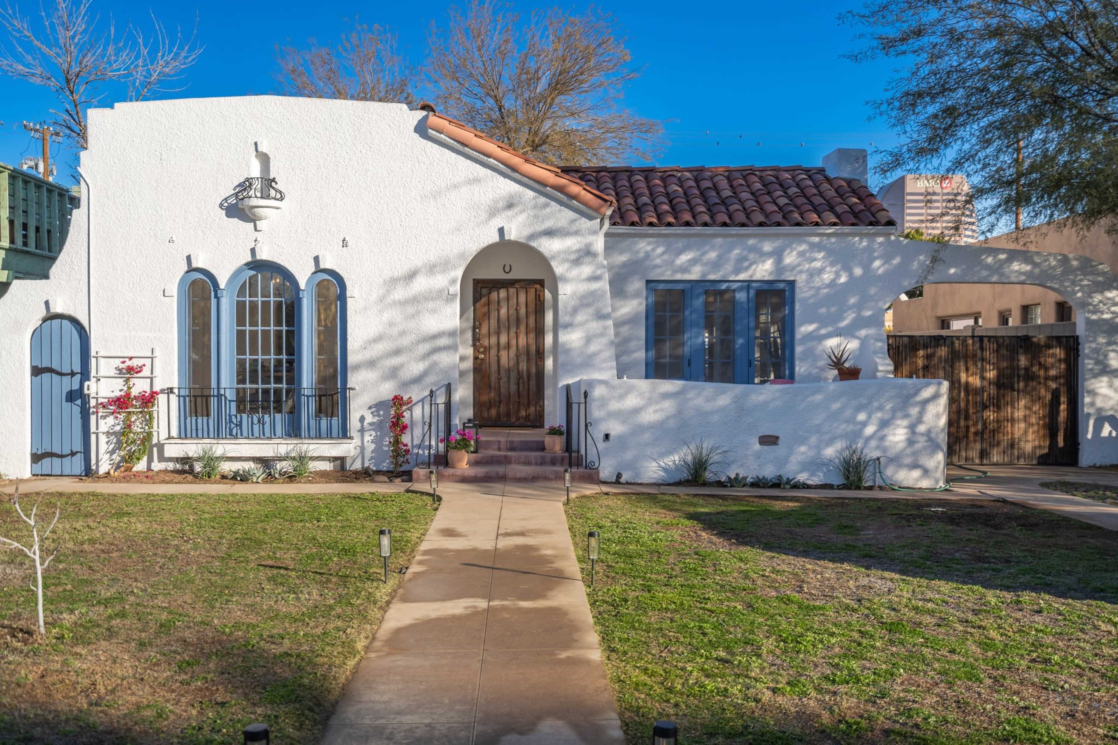 The image shows a white stucco house with a terracotta tiled roof, featuring arched windows and a wooden front door, surrounded by a manicured lawn and a concrete pathway.