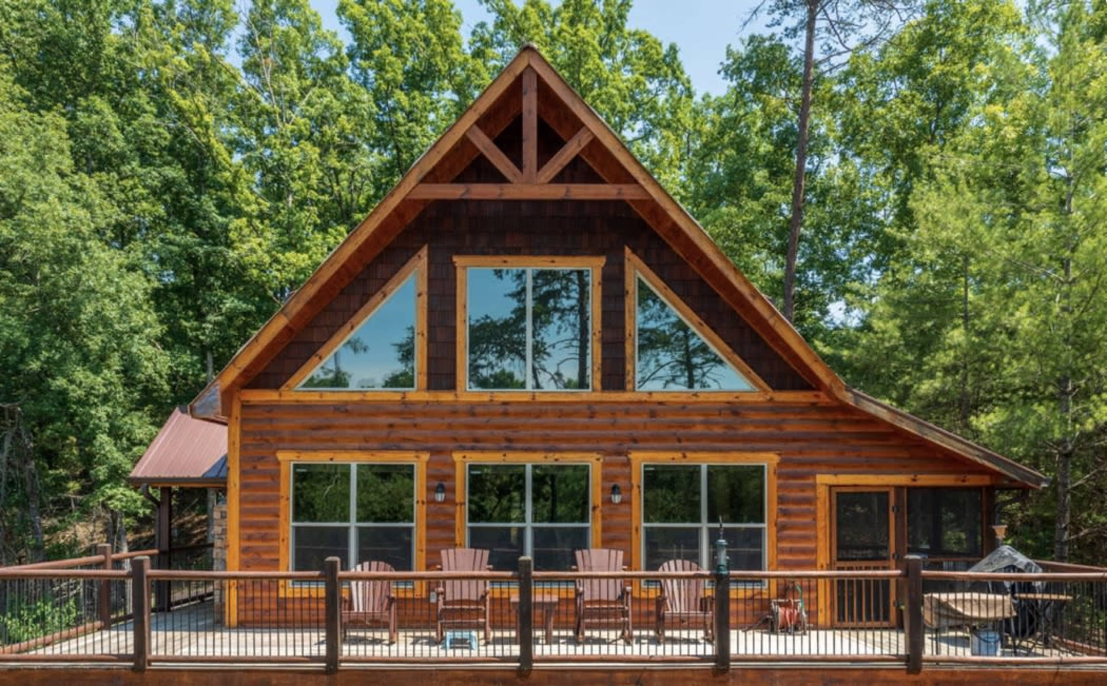 A wooden cabin with large windows and a deck featuring several chairs overlooks a dense forest.
