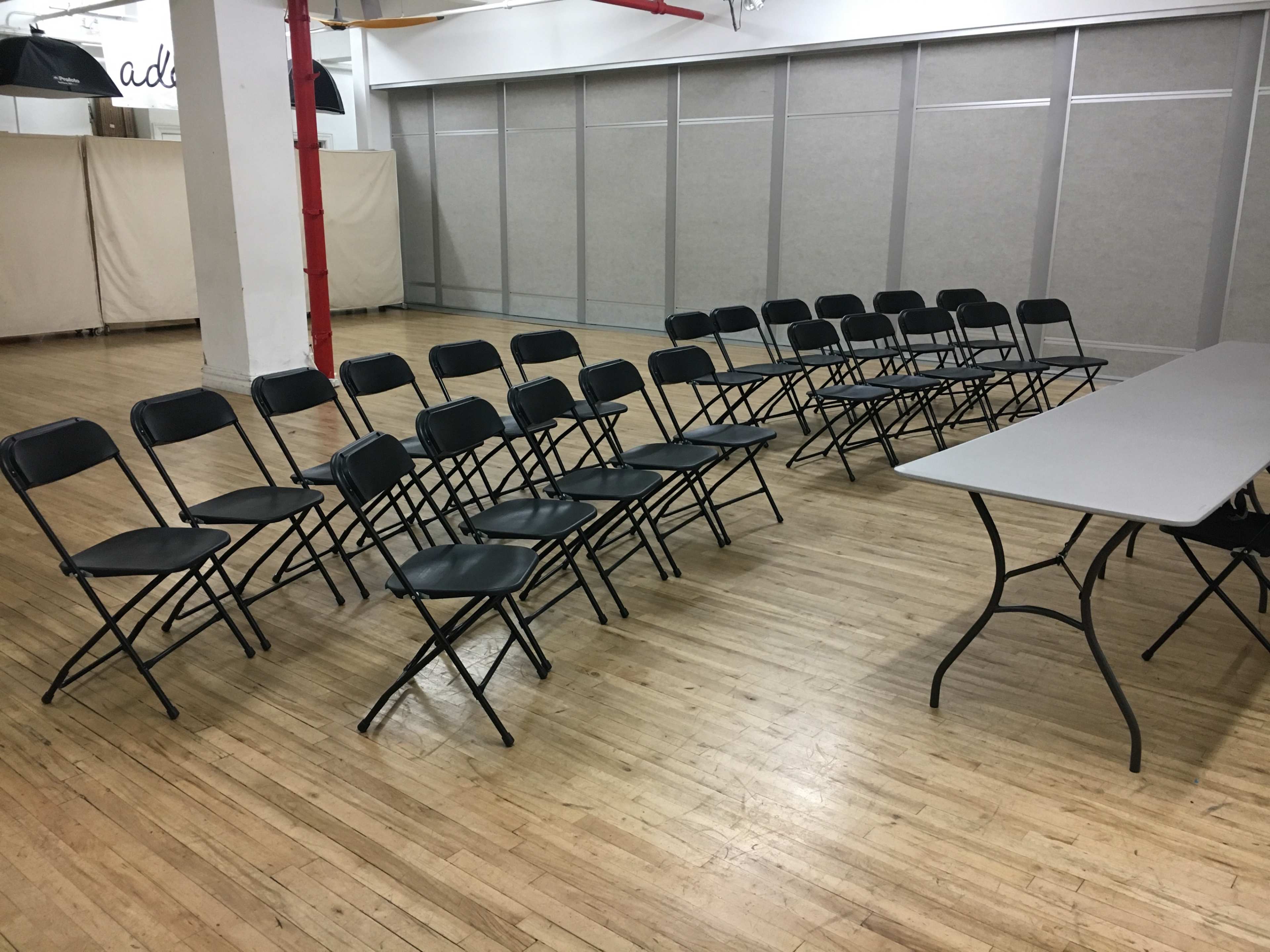 A set of black folding chairs is arranged in a semi-circle around a rectangular gray table in a spacious wooden-floored room.