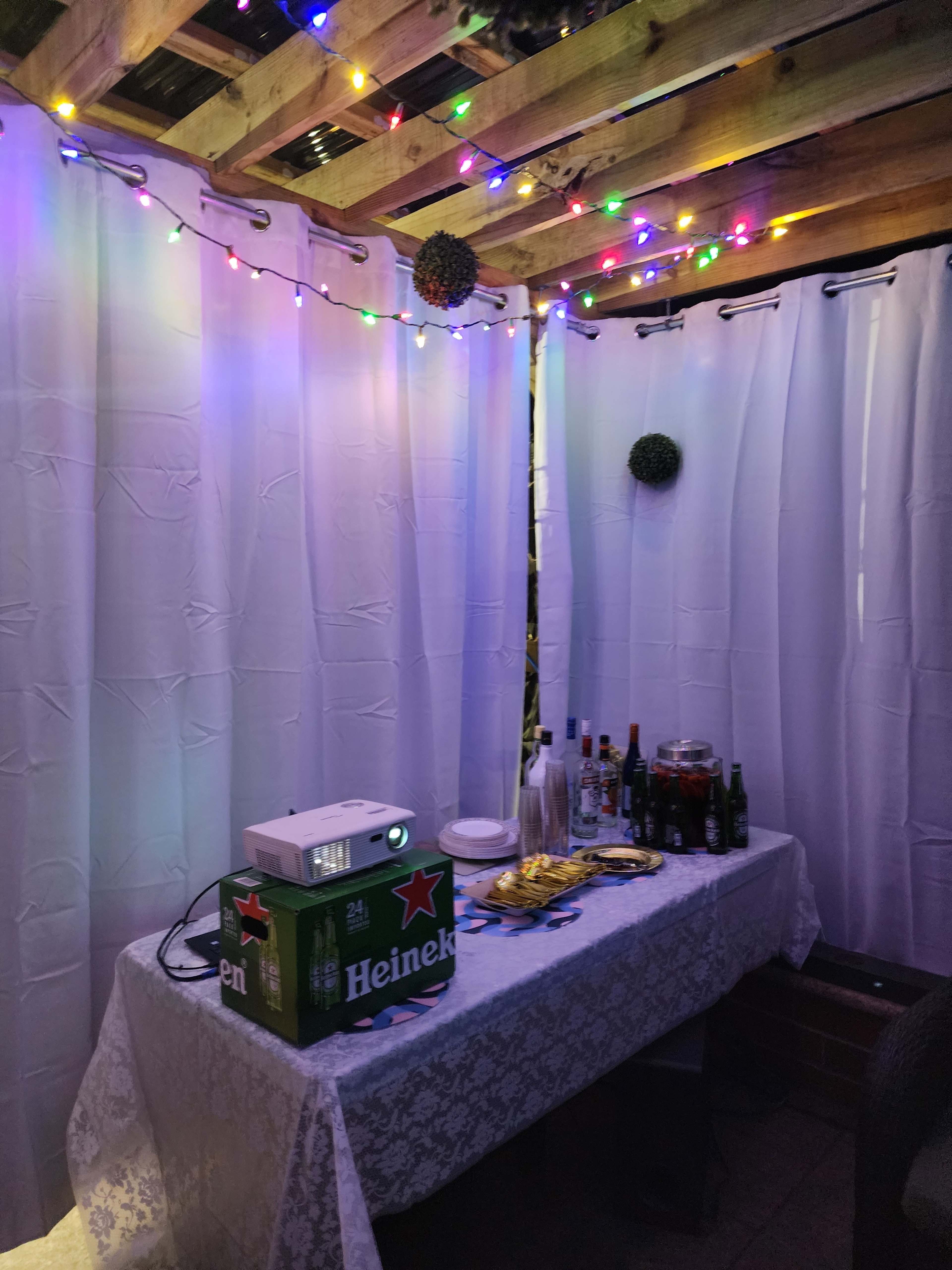 The scene features a table covered with a white cloth, adorned with various beverages, a projector, and surrounded by colorful string lights and curtains.