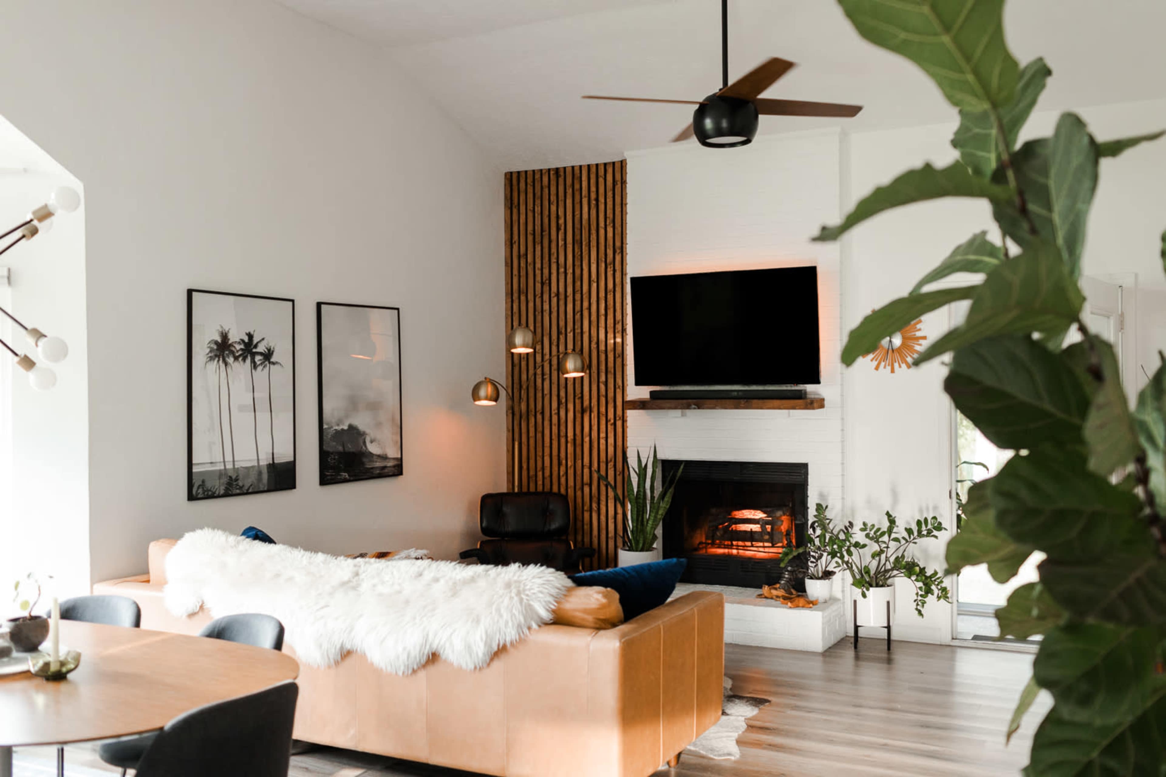 The image shows a modern living room with a leather couch, a wall-mounted TV, a fireplace, and framed photographs of palm trees.