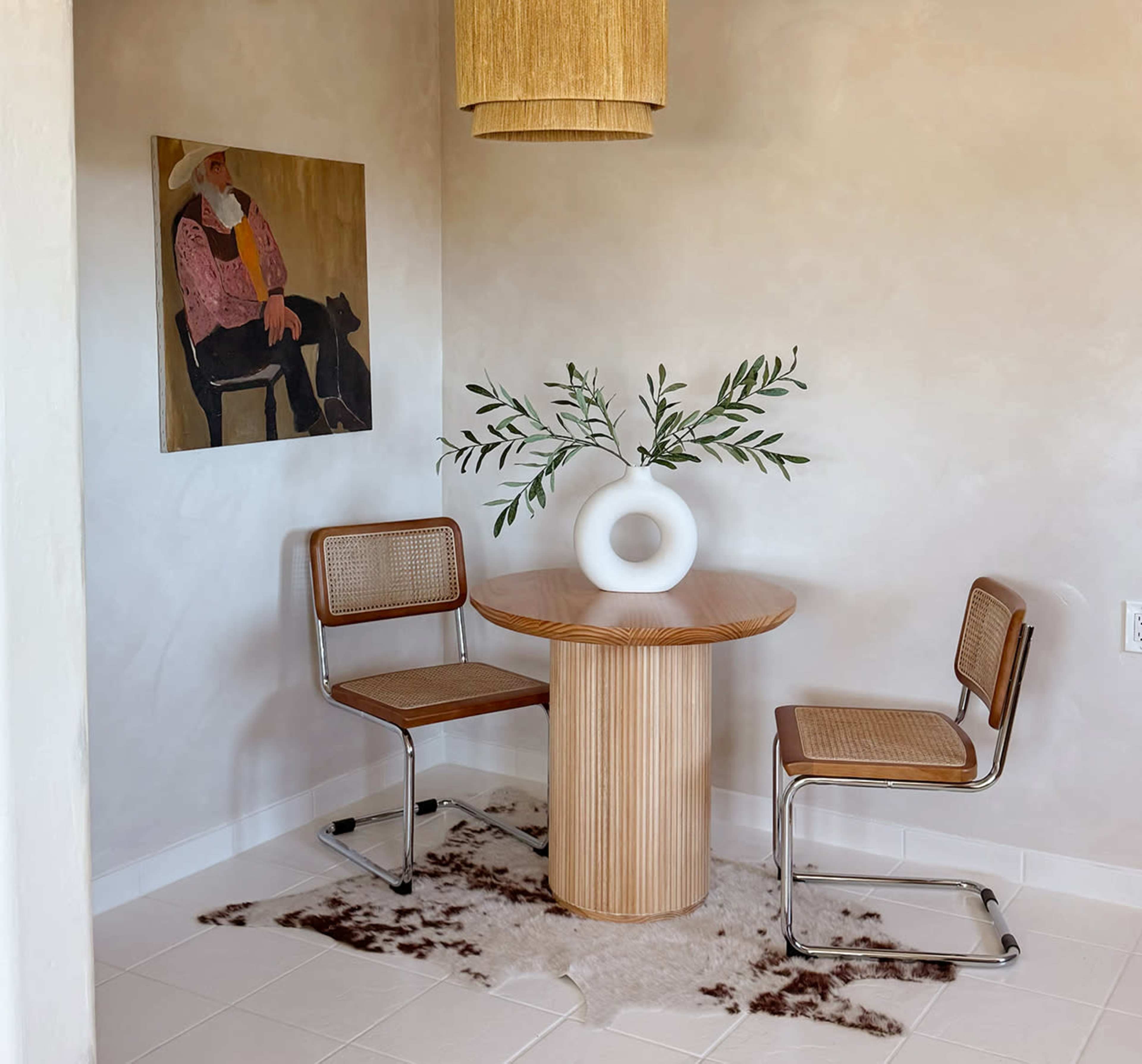 A round wooden table with two rattan chairs is set against a wall featuring an artwork of a man and a dog, with a hanging light and a vase holding greenery.