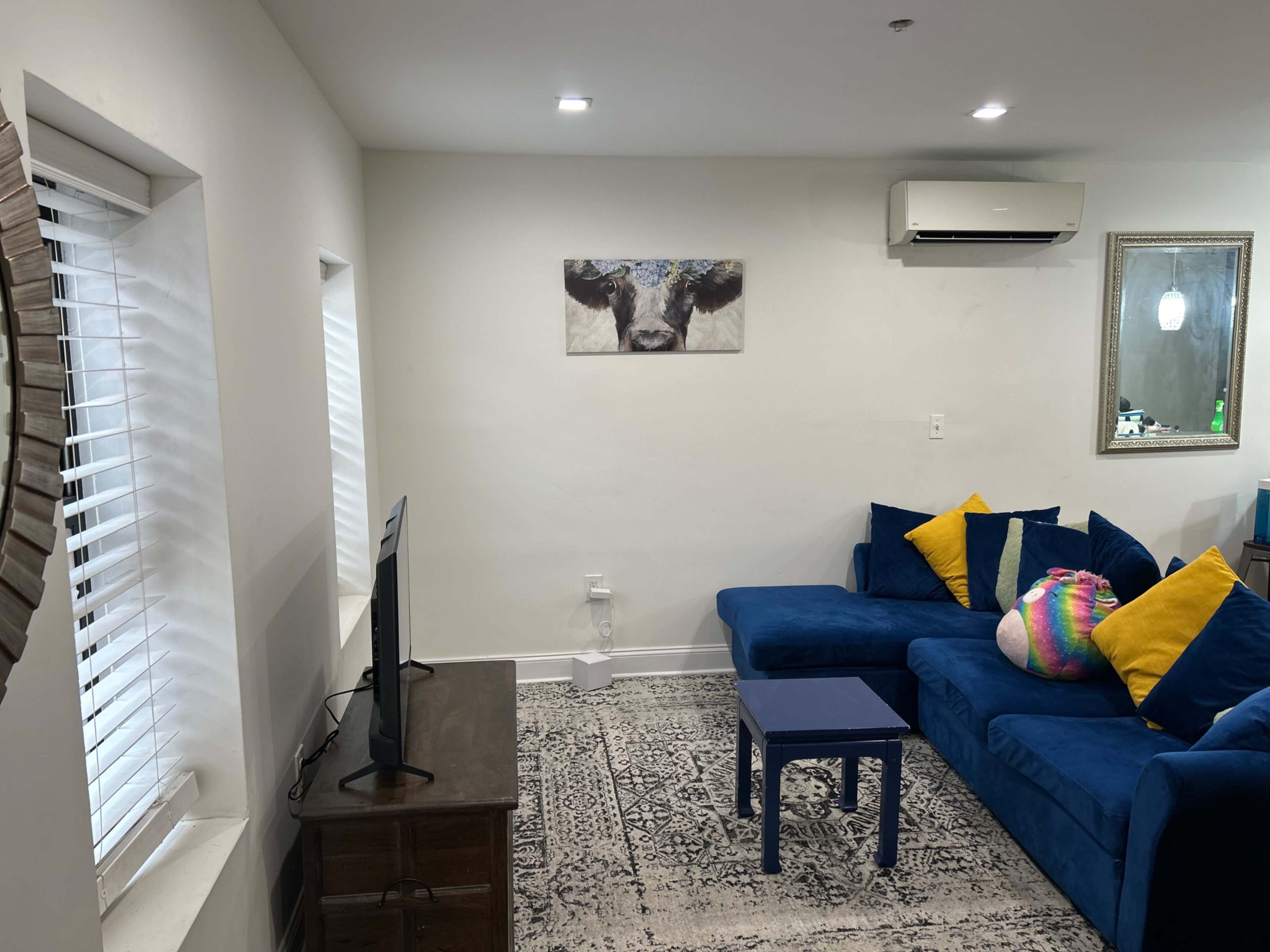 The image shows a living room with a blue sofa adorned with yellow pillows, a blue table, a television, and a cow-themed artwork on the wall.