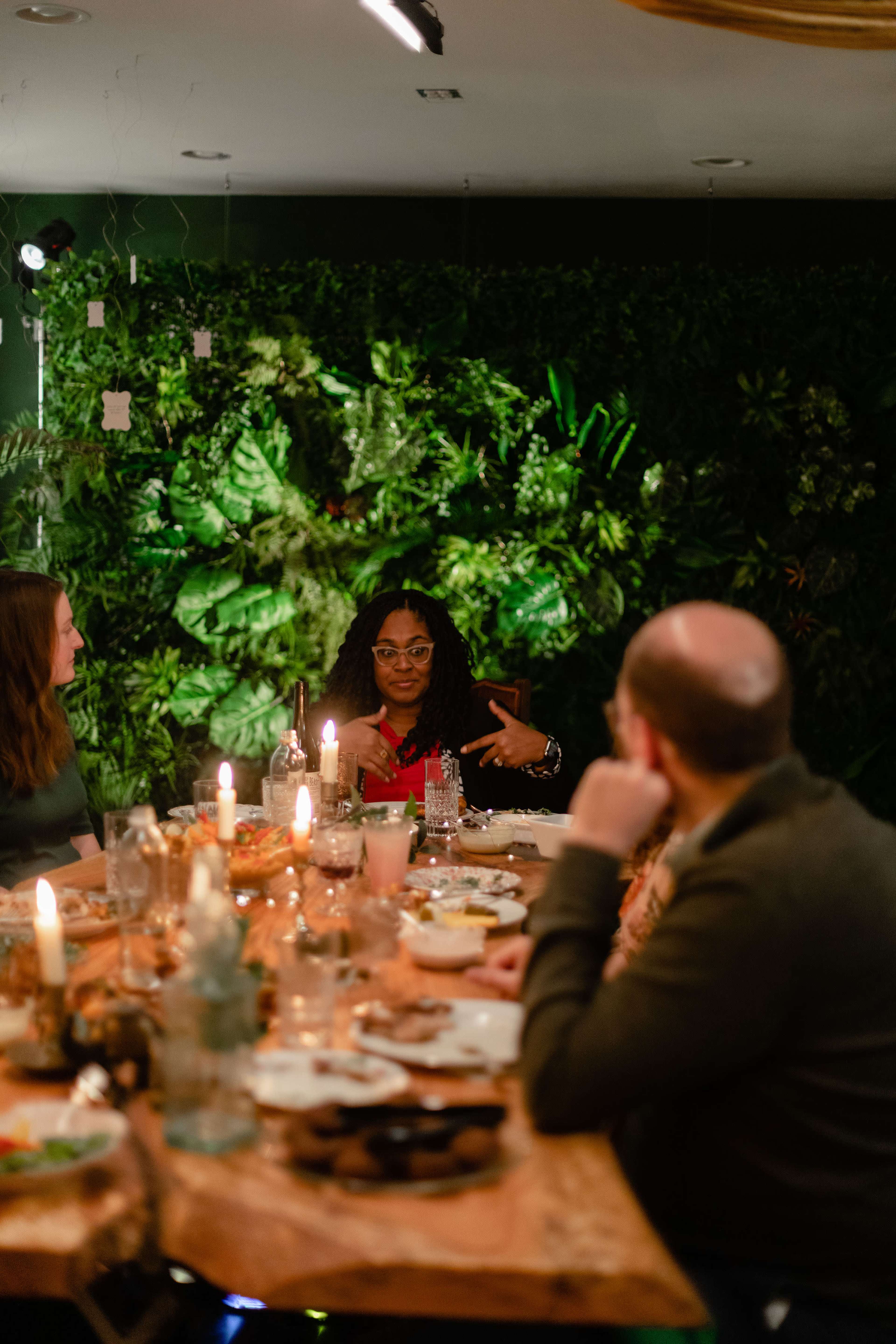 A group of people is seated around a table with candles, engaged in conversation amidst a backdrop of lush green plants.