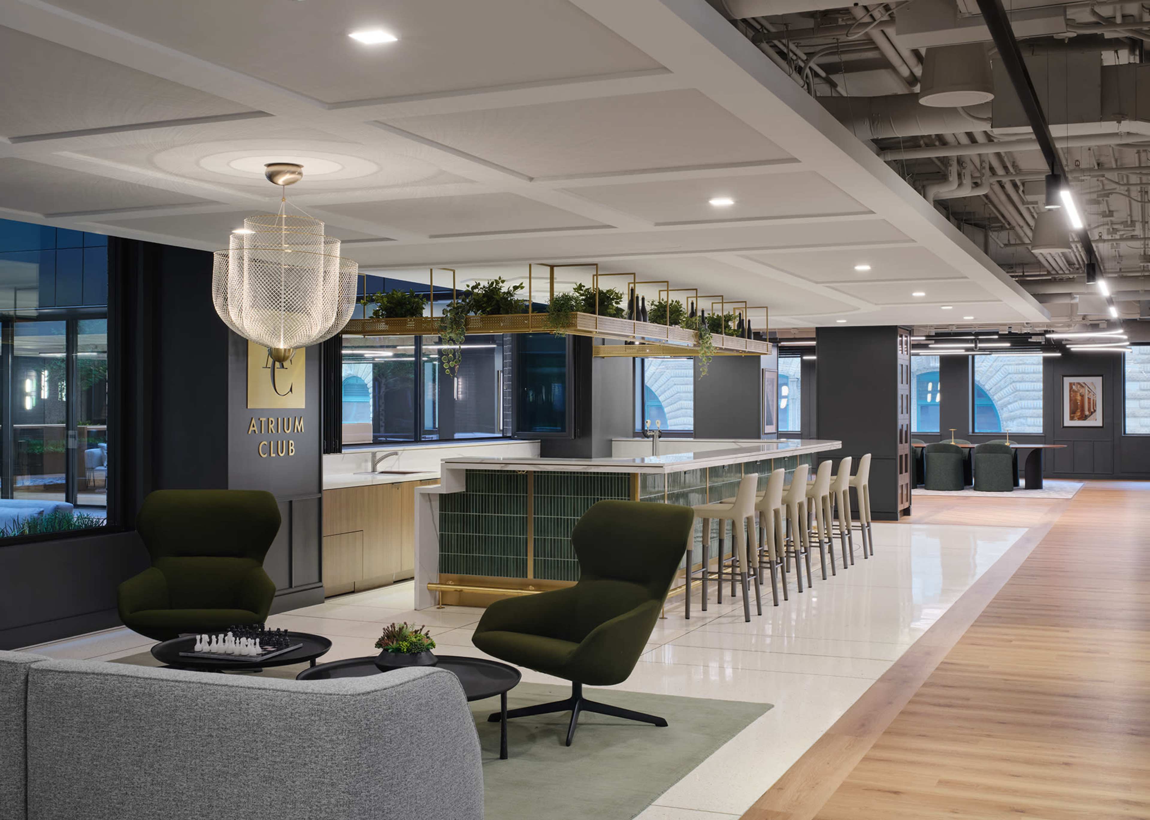 The image shows a modern office lounge area featuring a bar with high stools, green plants, contemporary furniture, and ample natural light from large windows.