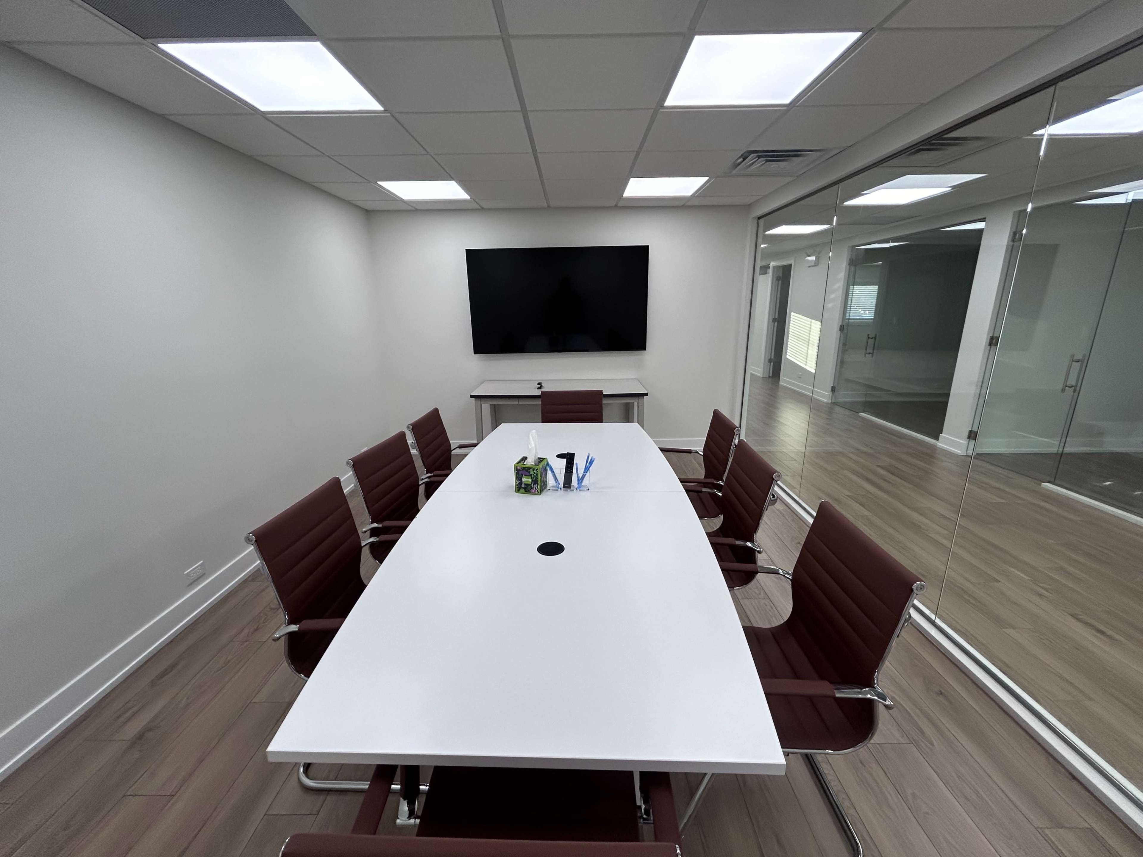 A modern conference room features a large white table surrounded by six brown leather chairs, with a flat-screen TV mounted on the wall and glass panels on one side.
