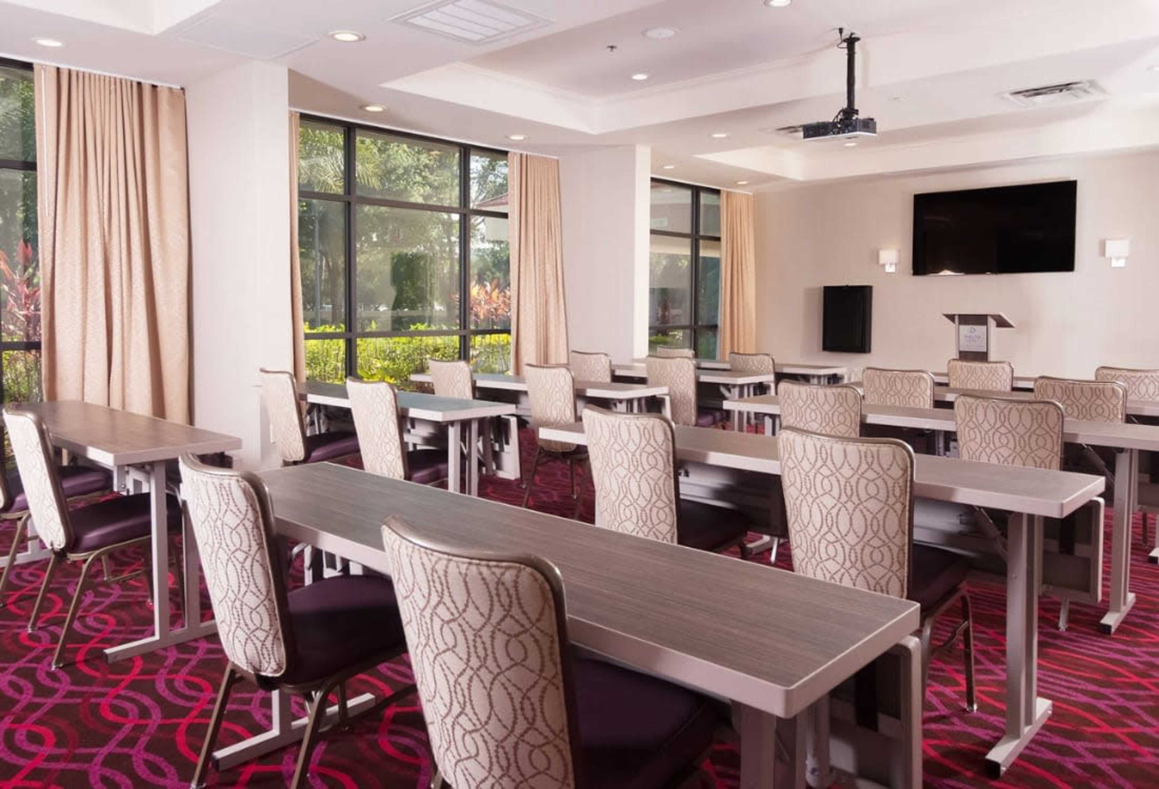 The image shows a conference room with multiple rows of tables and chairs arranged neatly, large windows allowing natural light, and a projector screen on the front wall.