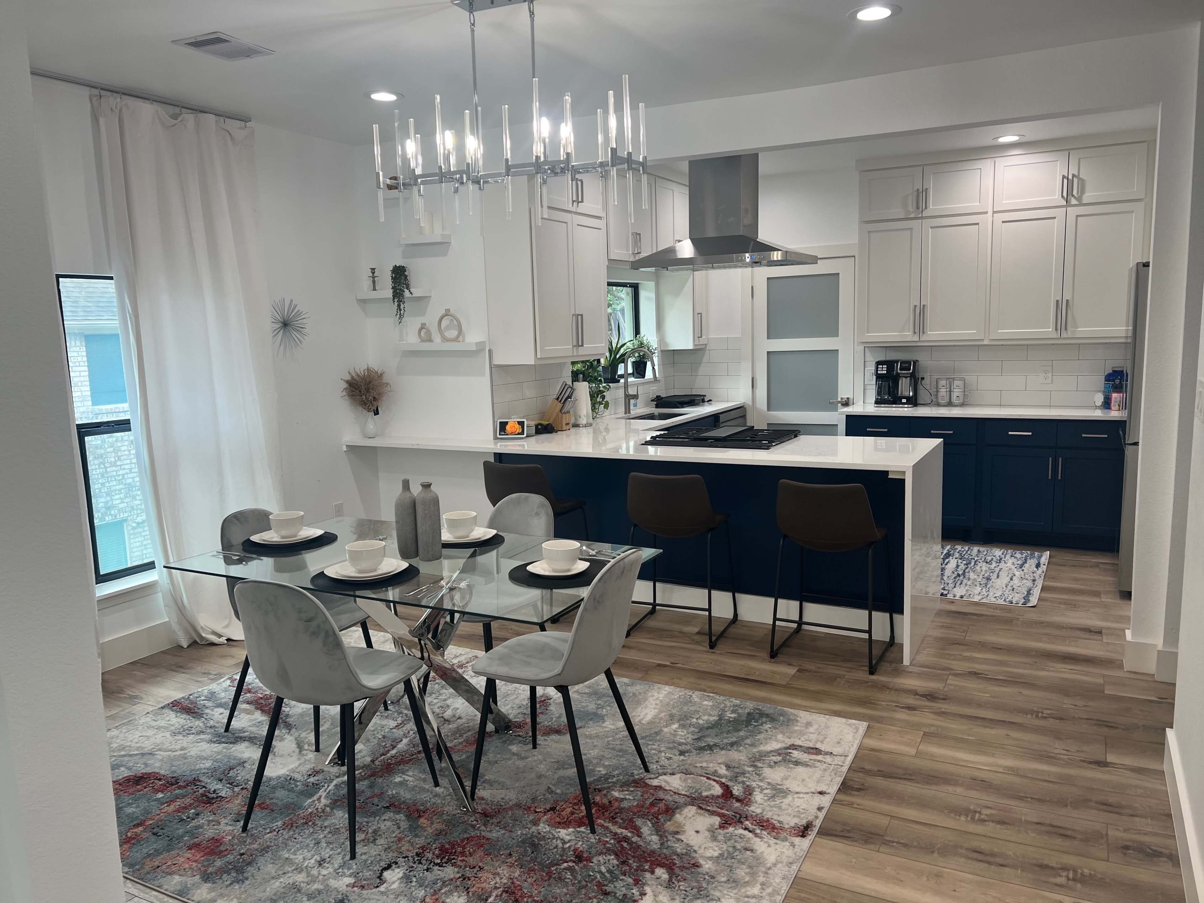 A modern kitchen and dining area feature a glass table set for dinner, surrounded by stylish chairs and illuminated by a chandelier.