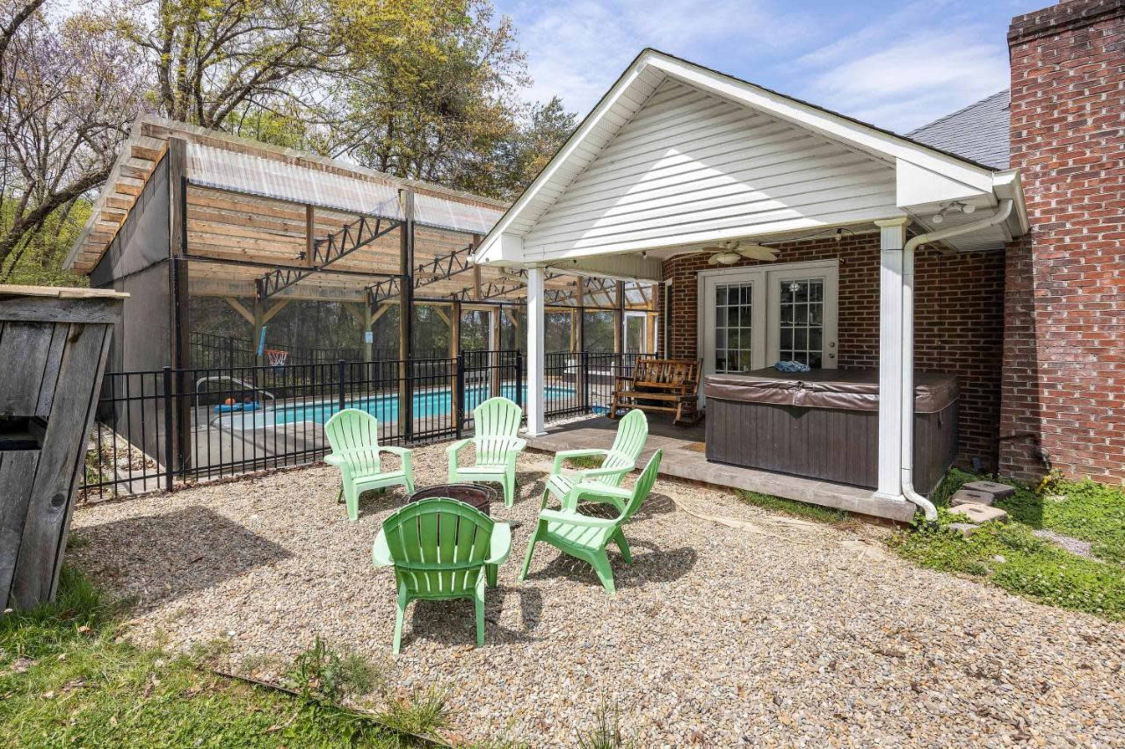 A backyard area features green adirondack chairs set in a gravel space near a covered hot tub and a screened-in pool.