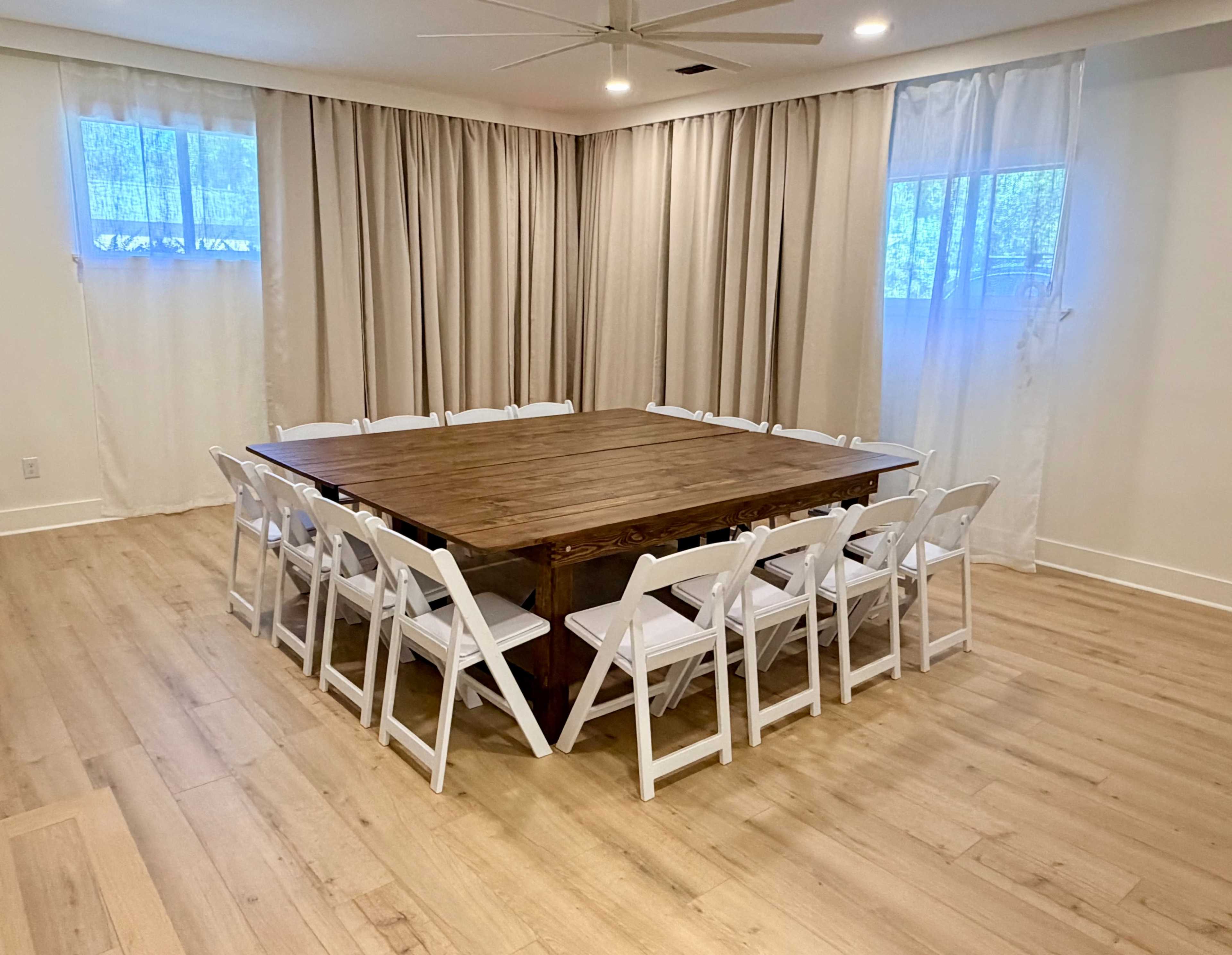 A large wooden table surrounded by twelve white folding chairs is positioned in a well-lit room with light curtains and hardwood flooring.