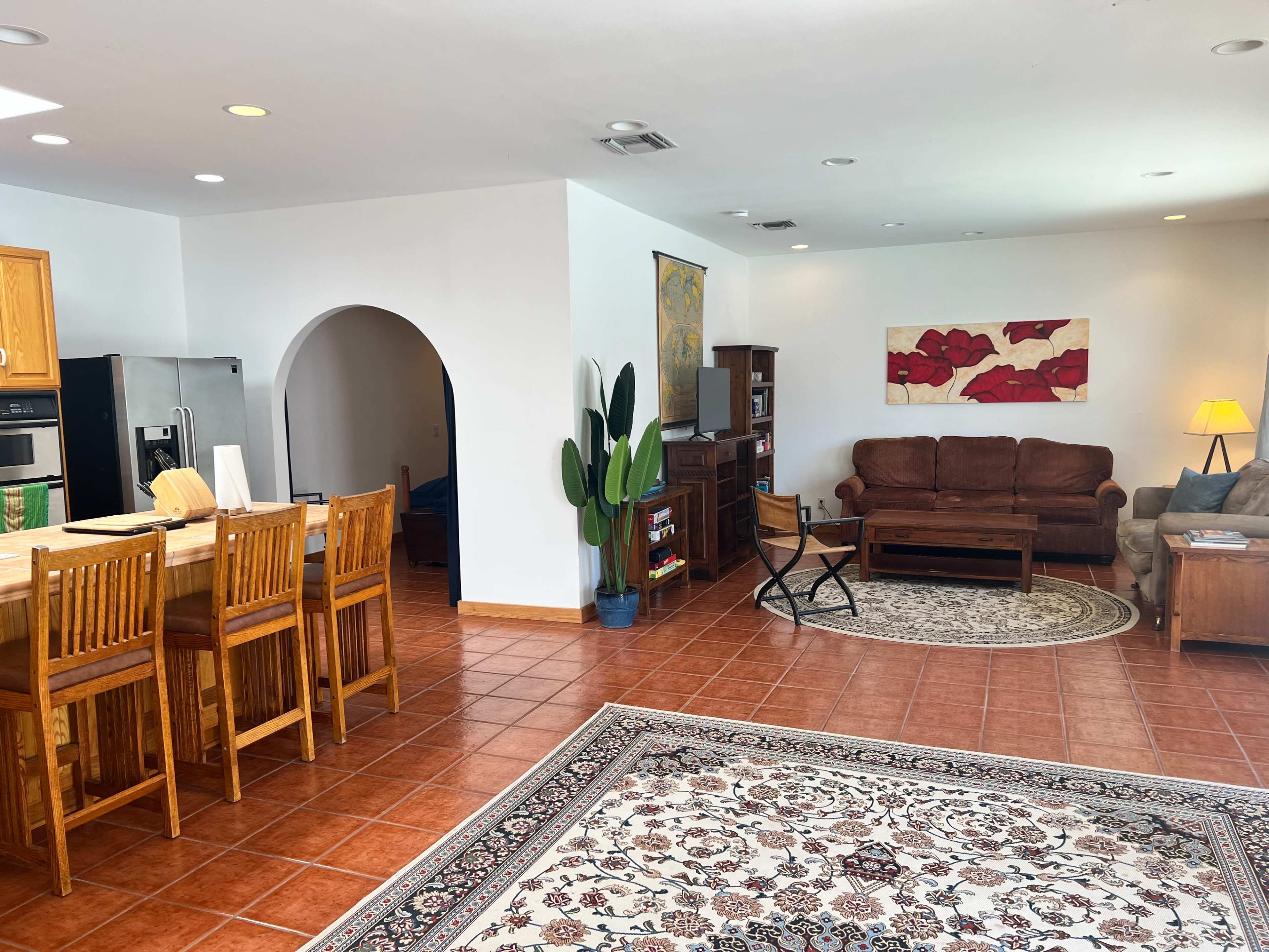 The image shows a spacious living room and kitchen area, featuring wooden bar stools at a counter, a comfortable seating arrangement, and a large rug on the tiled floor.