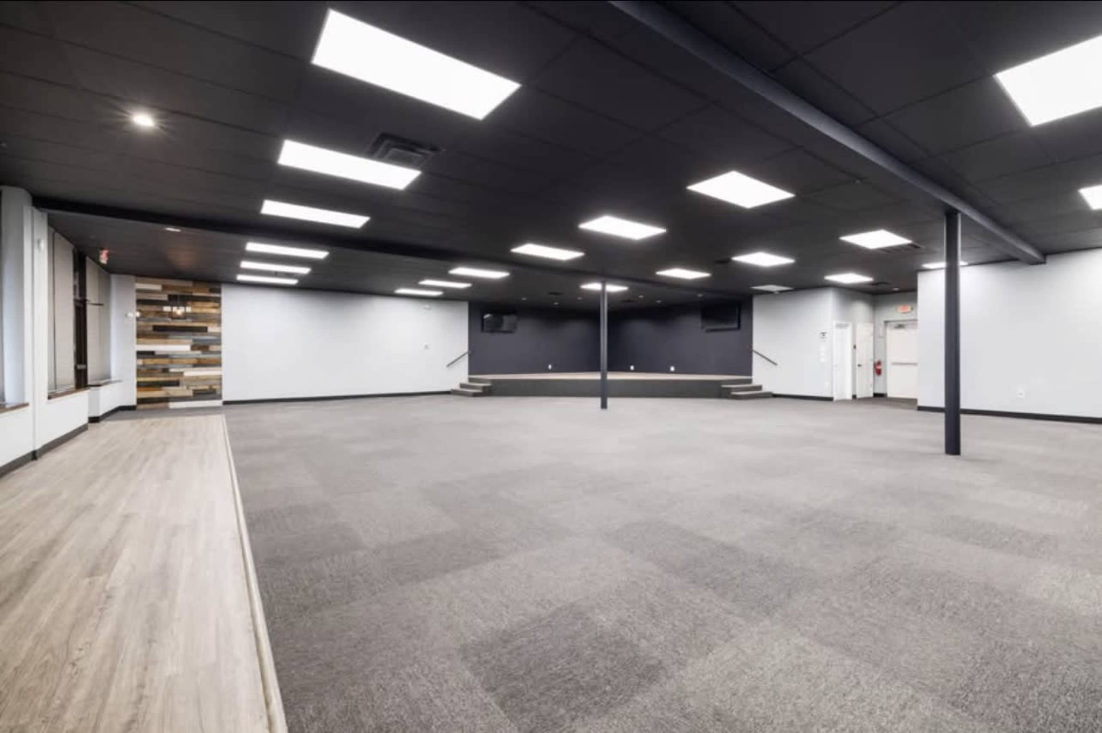 The image shows a spacious, empty room with gray carpeting, a dark wall at one end, and fluorescent ceiling lights.