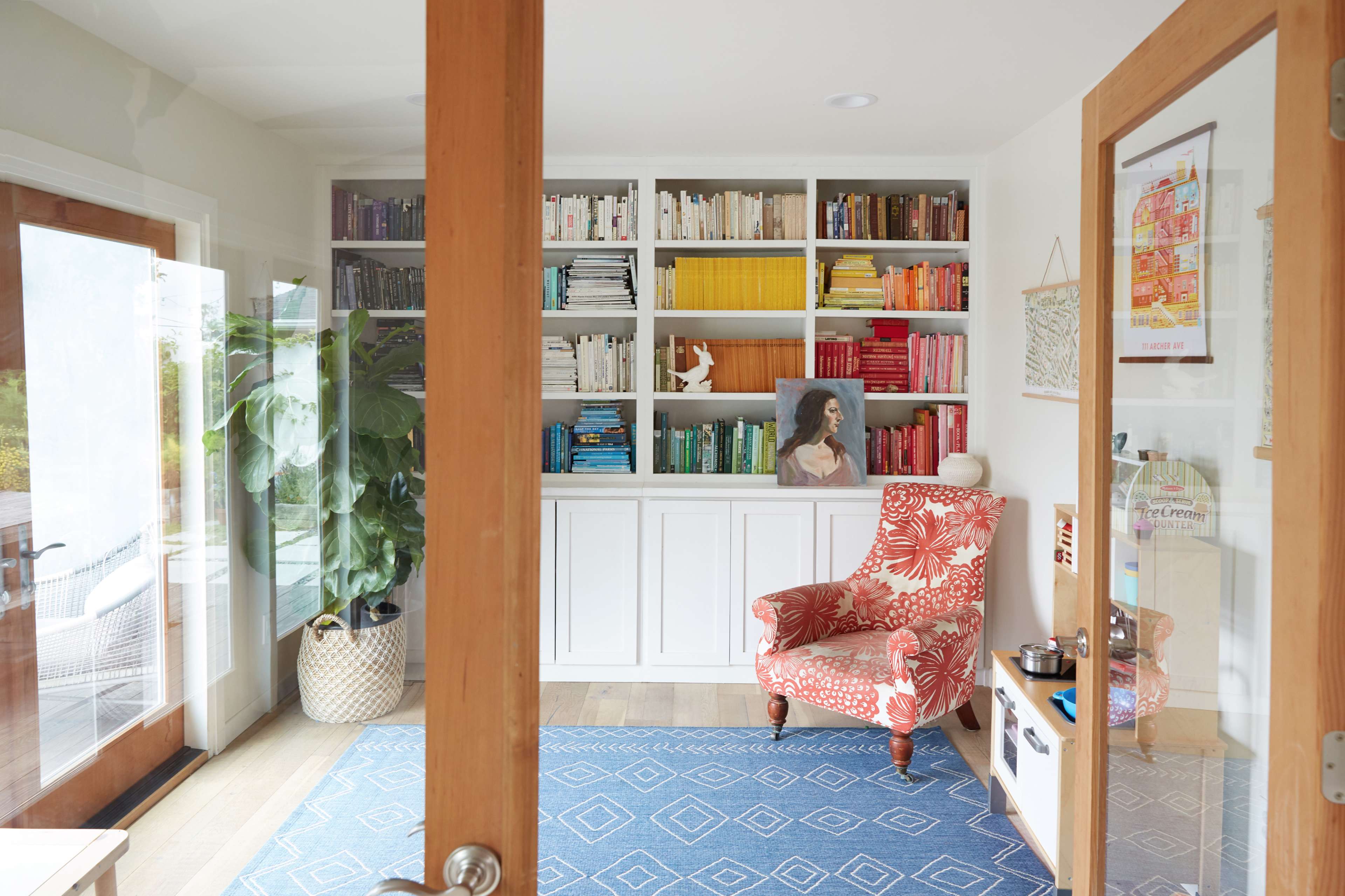 A cozy reading nook features a patterned chair beside a bookshelf filled with colorful books, framed by large glass doors and indoor plants.