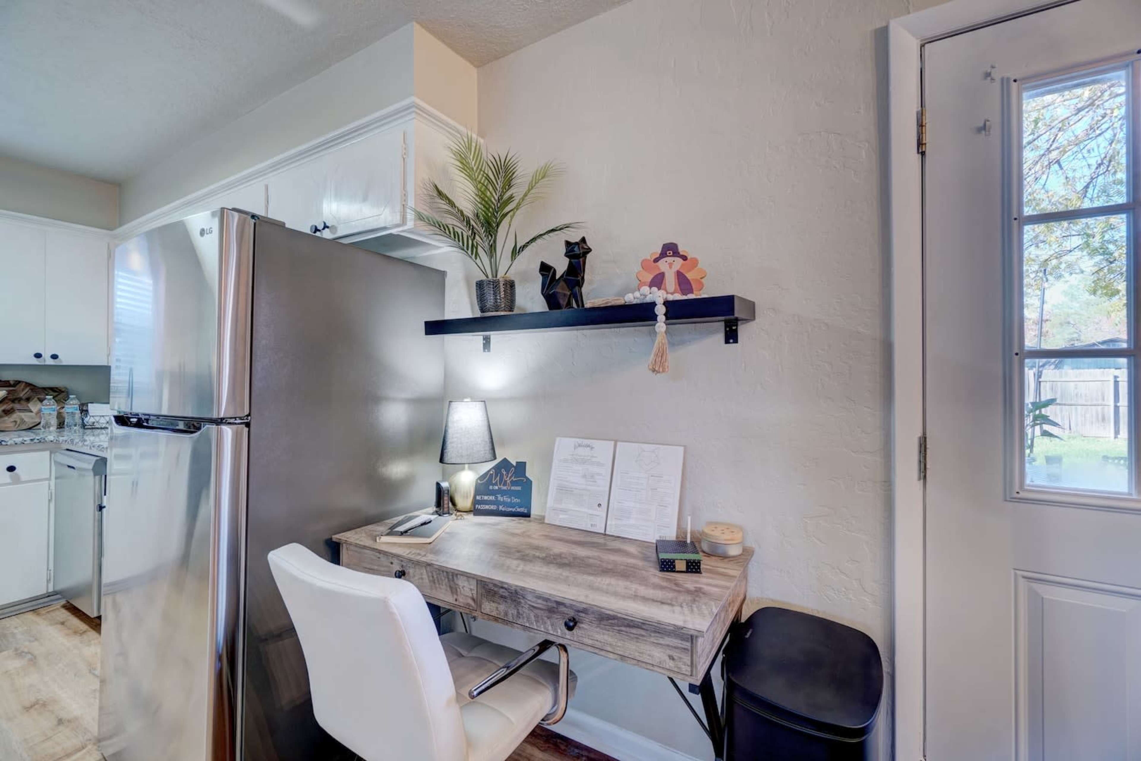 A small workspace with a desk and chair, positioned next to a refrigerator and near a door leading outside.