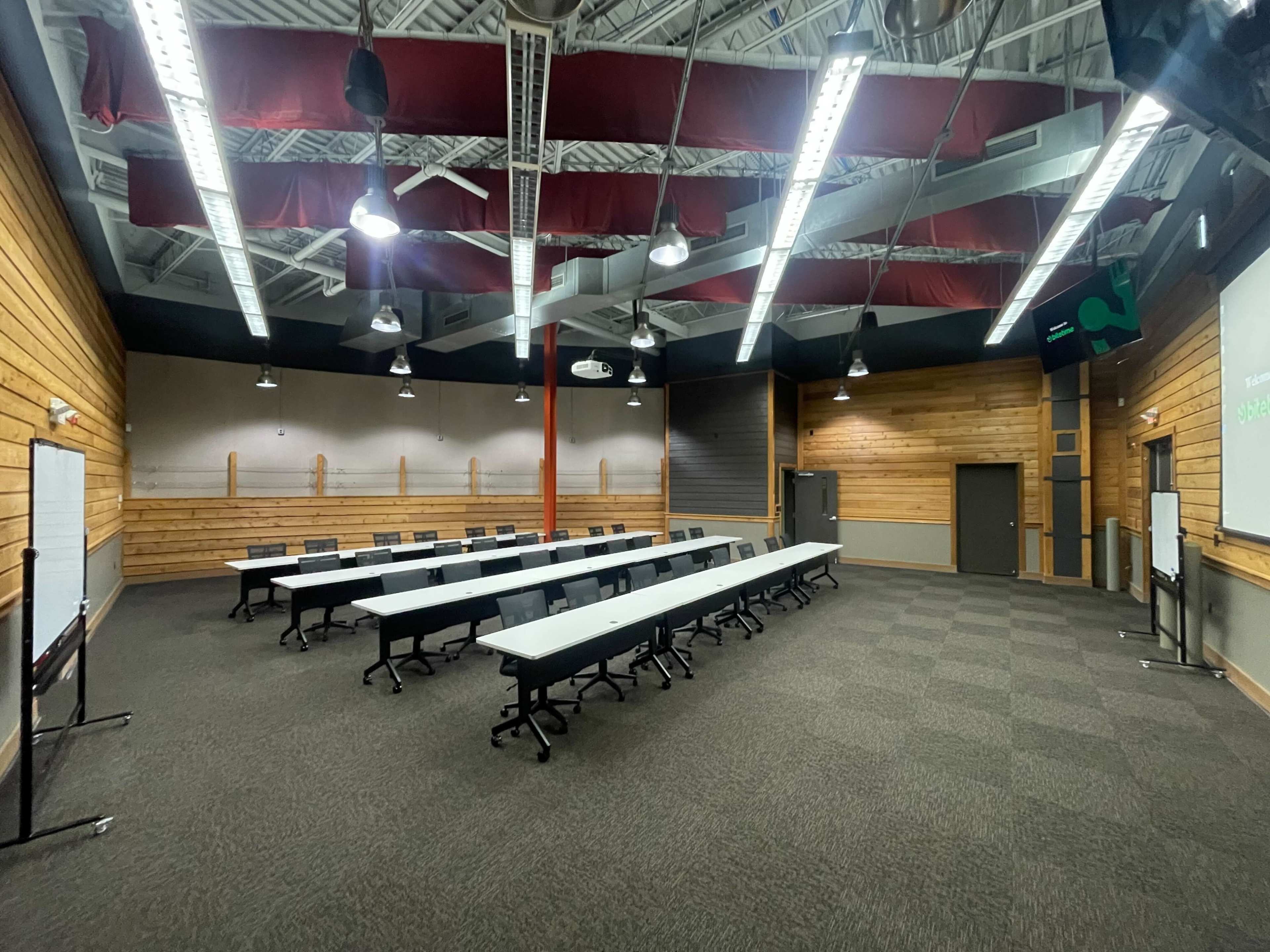 The image shows a spacious meeting room set up with multiple rectangular tables arranged in a U-shape, surrounded by wooden walls and equipped with overhead lighting.