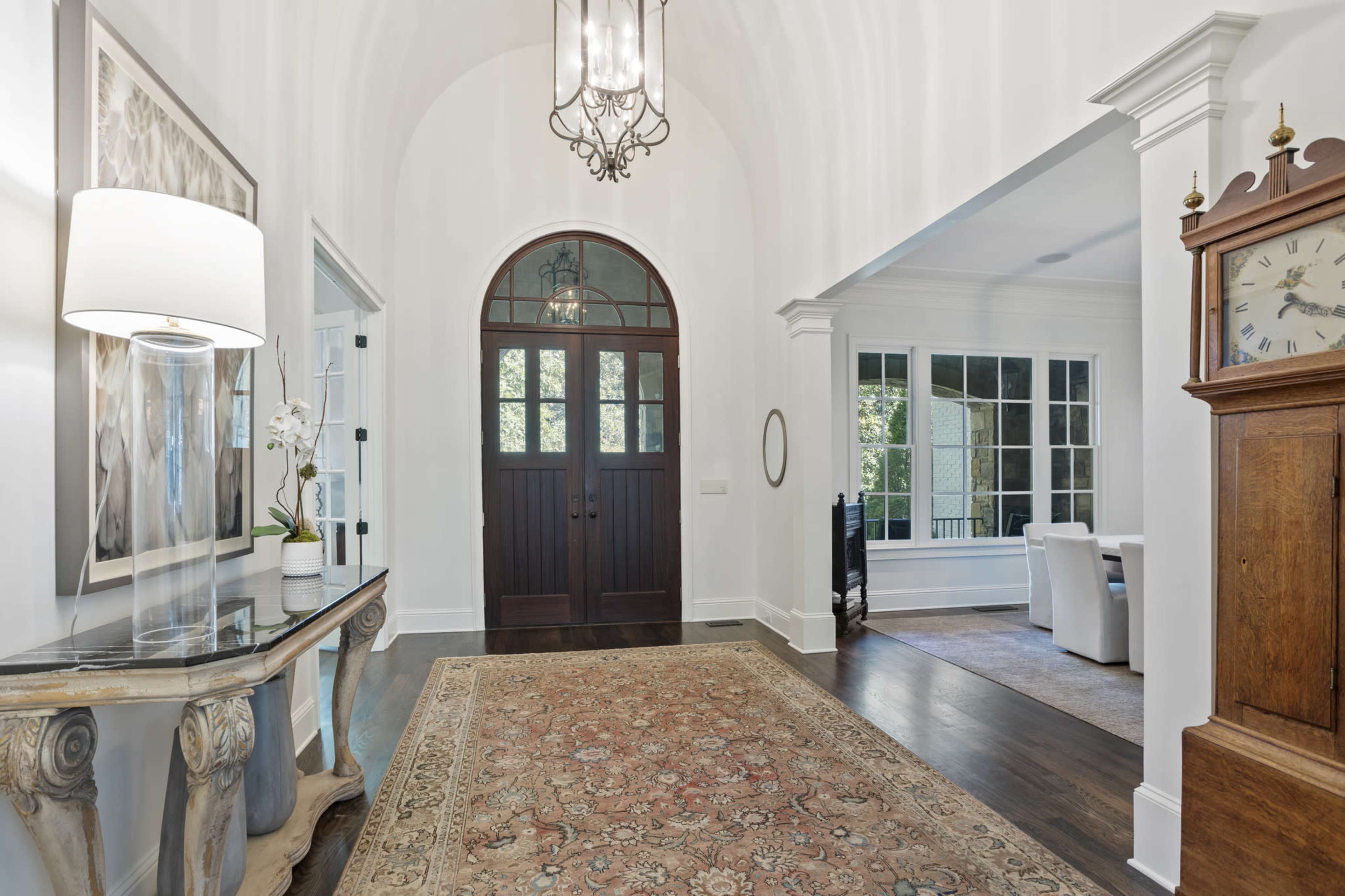A bright entryway with a large arched wooden door, a decorative table with a lamp, and a view of a living area through the doorway.