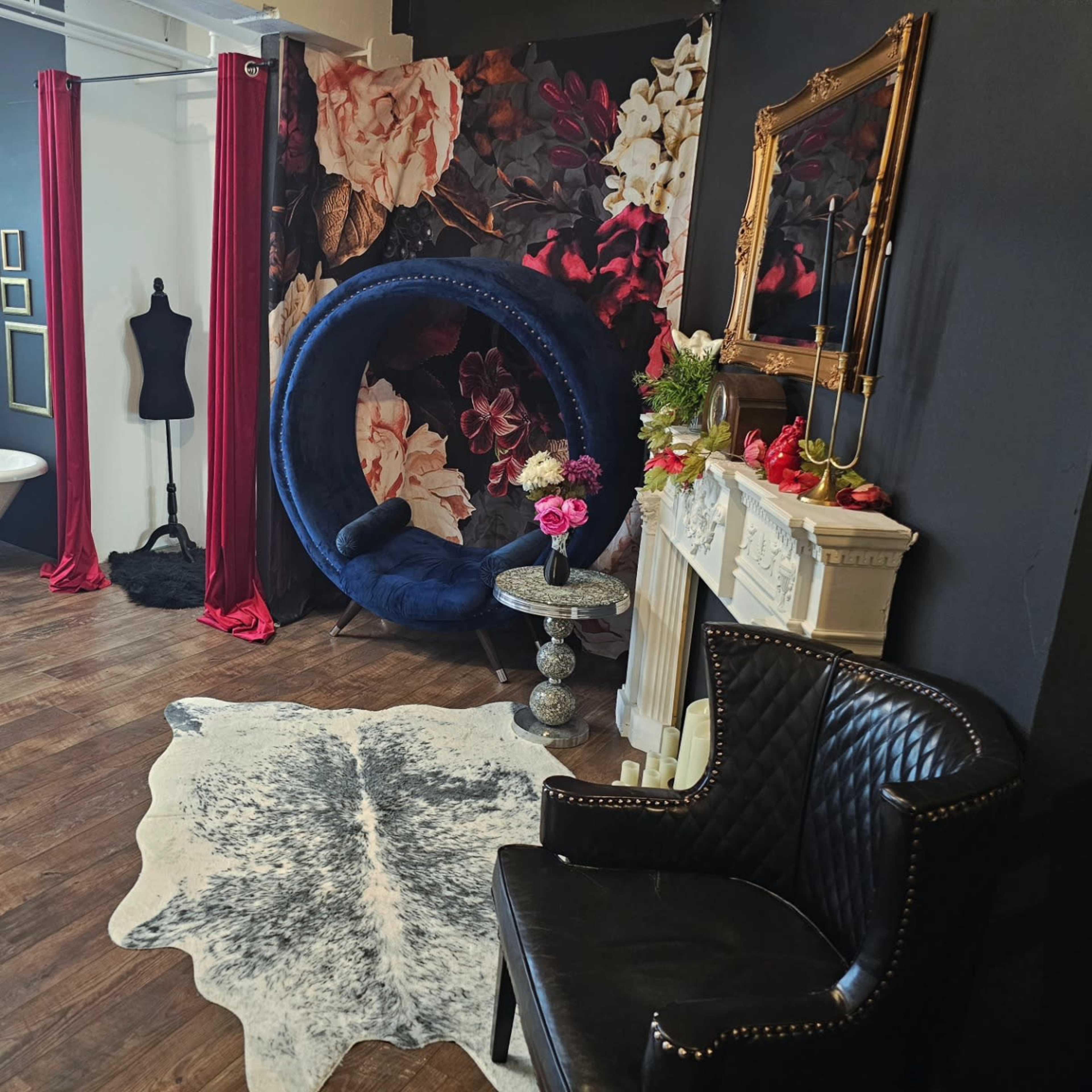 The scene features a stylish interior with a floral backdrop, a blue circular chair, a black leather armchair, and decorative elements like a vintage mirror and flowers.