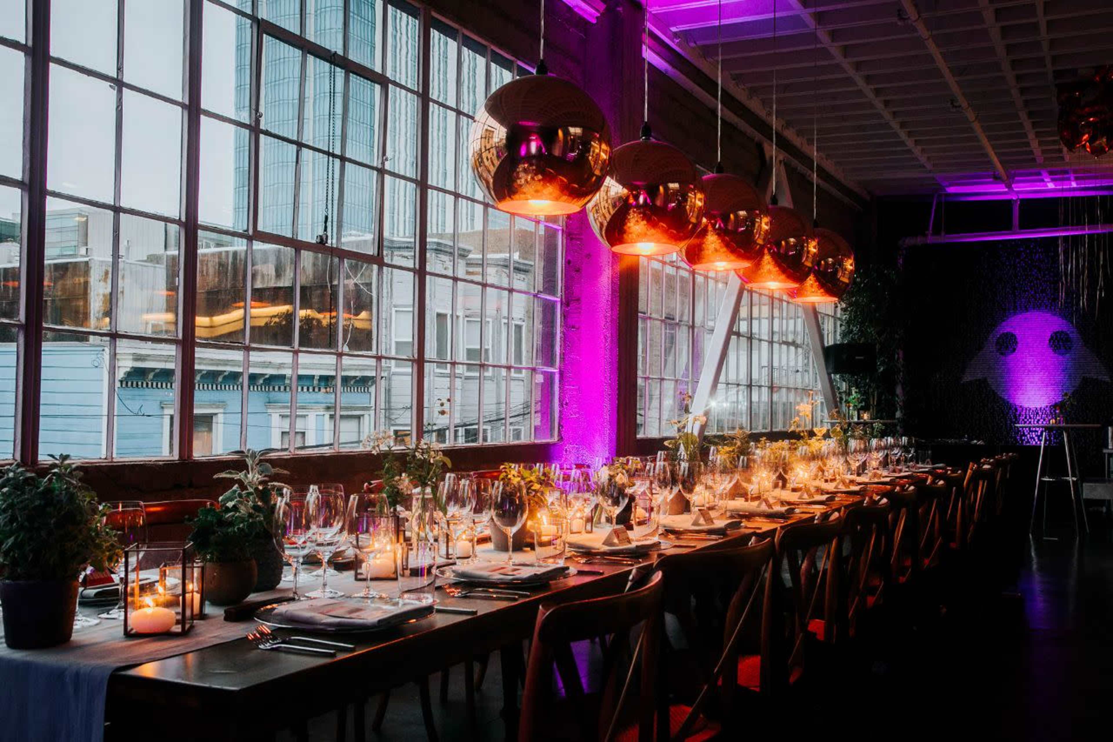 A long dining table is set with elegant dinnerware and surrounded by large windows, while warm pendant lights illuminate the space.