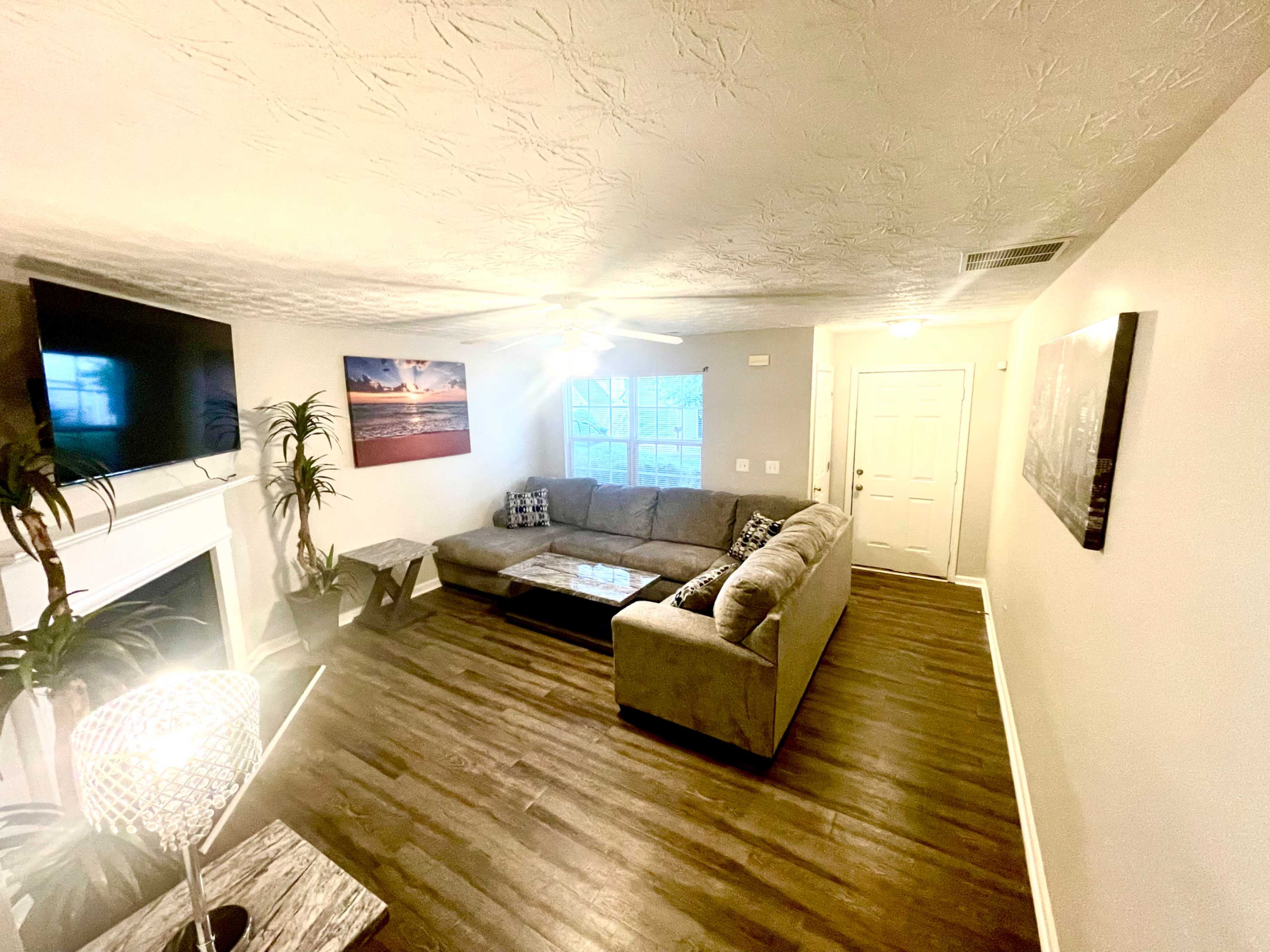 The living room features a sectional sofa, a coffee table, a television mounted on the wall, and a front door with natural light entering through a window.
