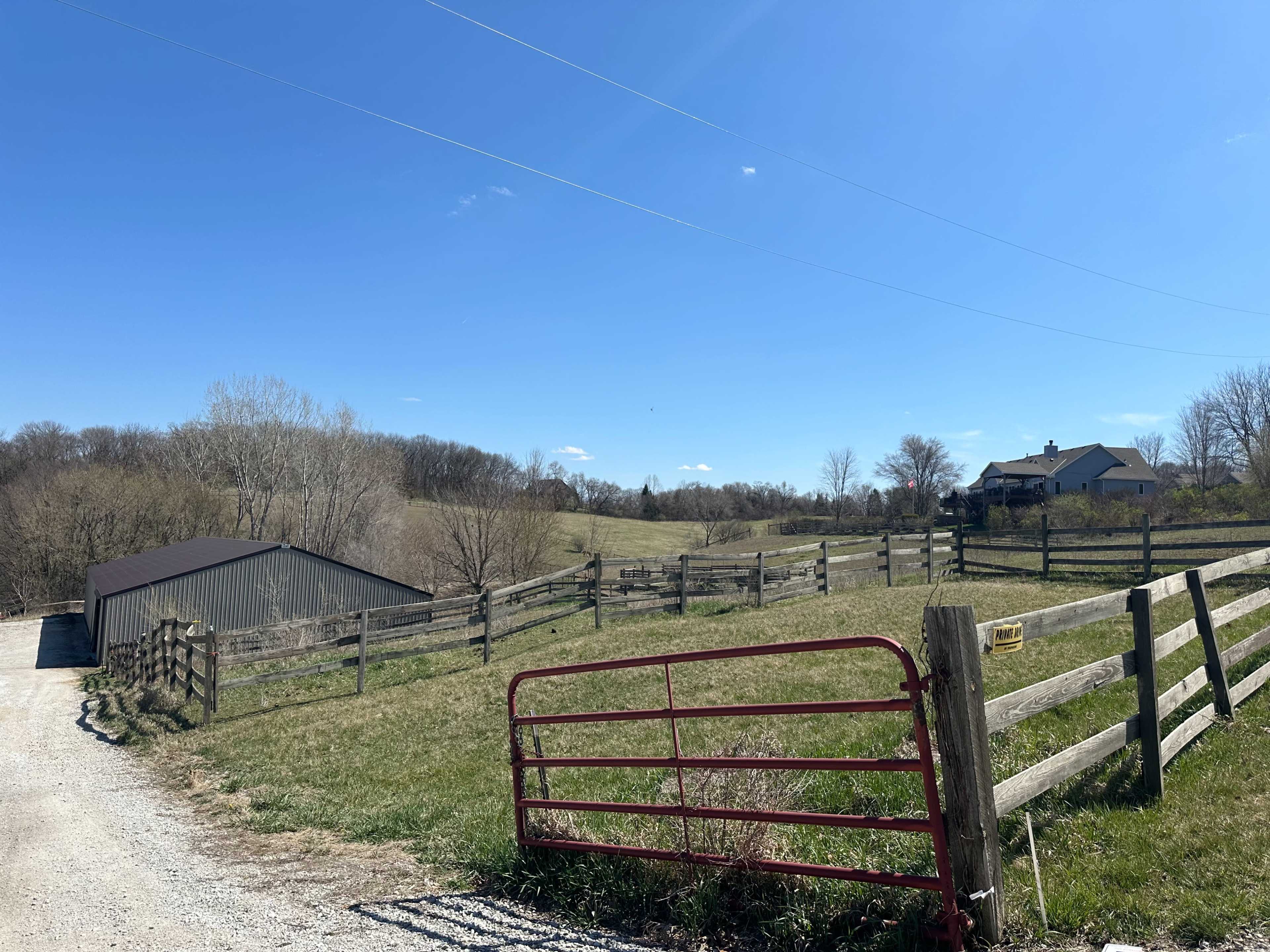 A rural landscape featuring a gravel driveway leading to a red gate, with a barn and house visible in the distance surrounded by wooden fences and trees.