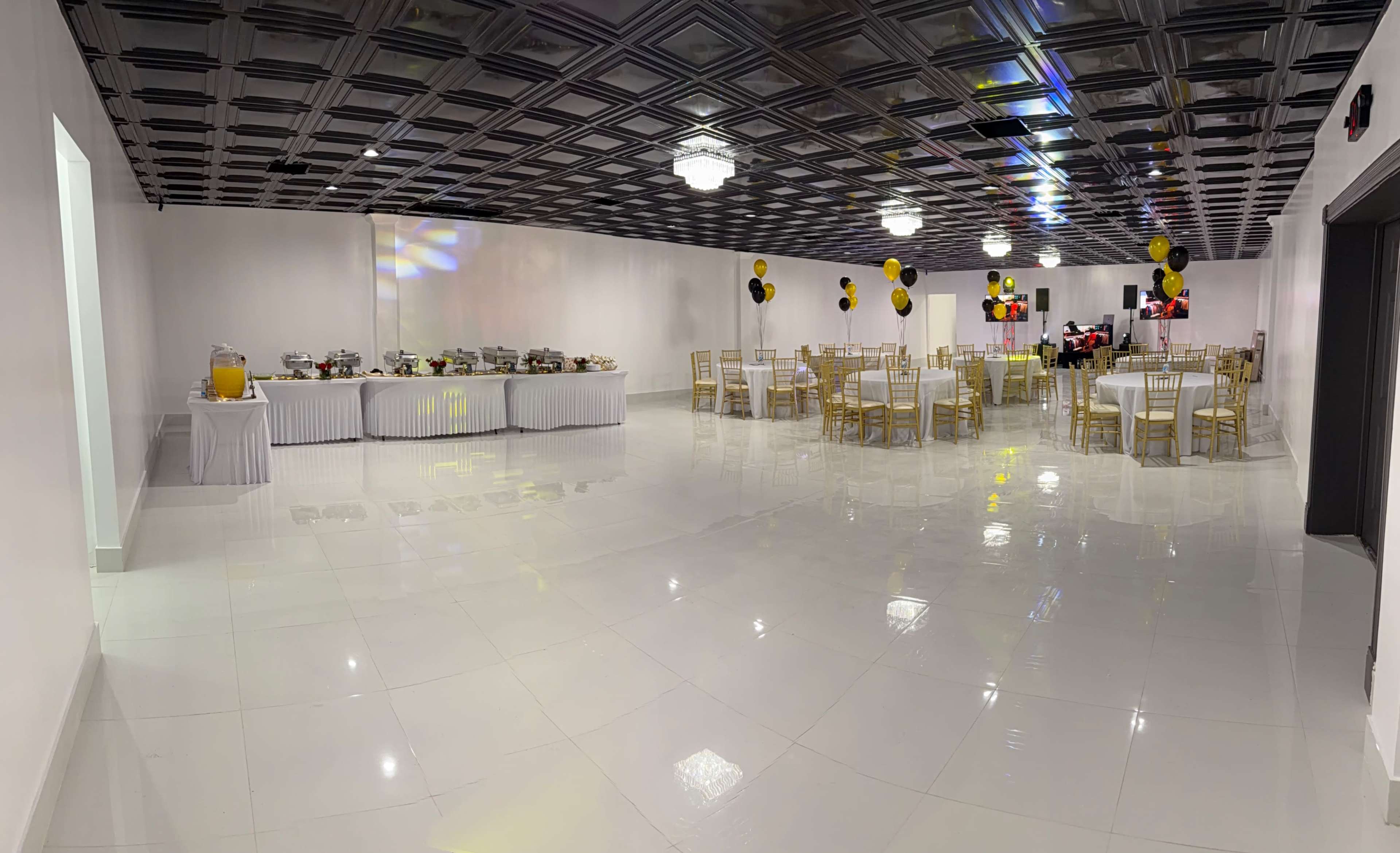 The image shows a spacious, elegantly arranged banquet hall with round tables, gold chairs, and a buffet table set against a white backdrop.
