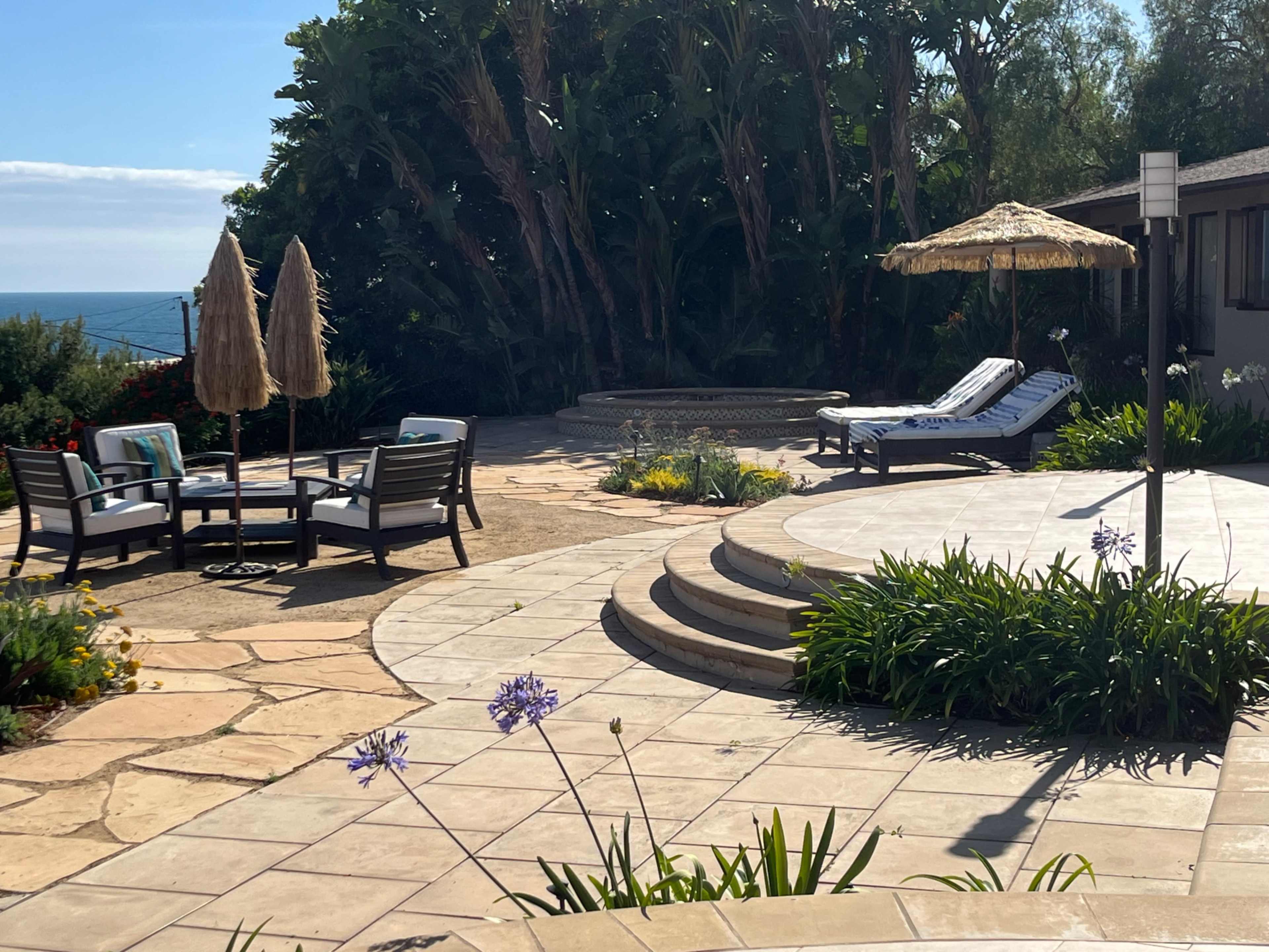The image shows a landscaped outdoor patio area with seating, a lounge chair, and a thatched umbrella overlooking the ocean.