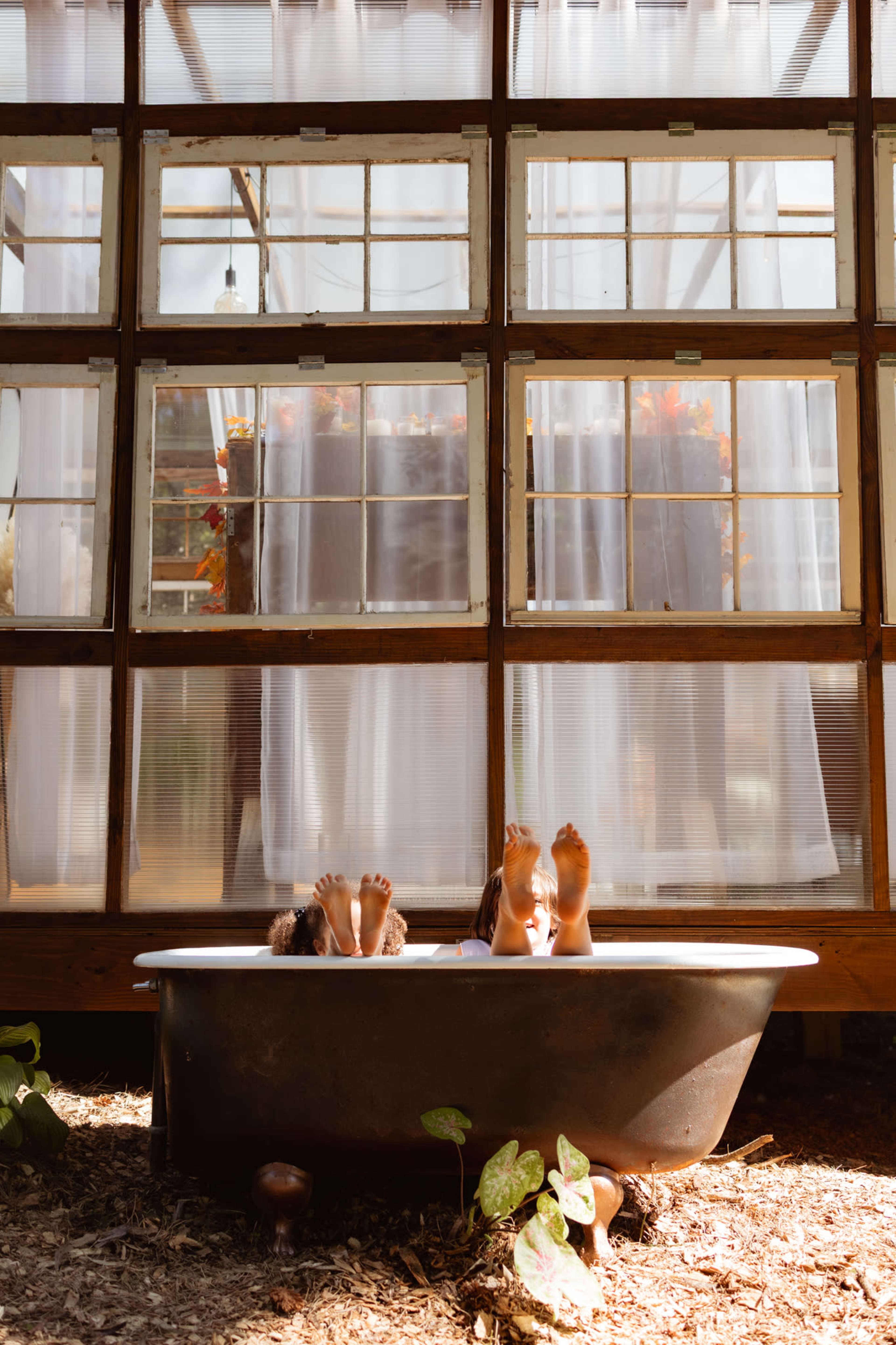 Two children sit in a vintage bathtub with their feet visible, positioned in front of a large window with sheer white curtains.