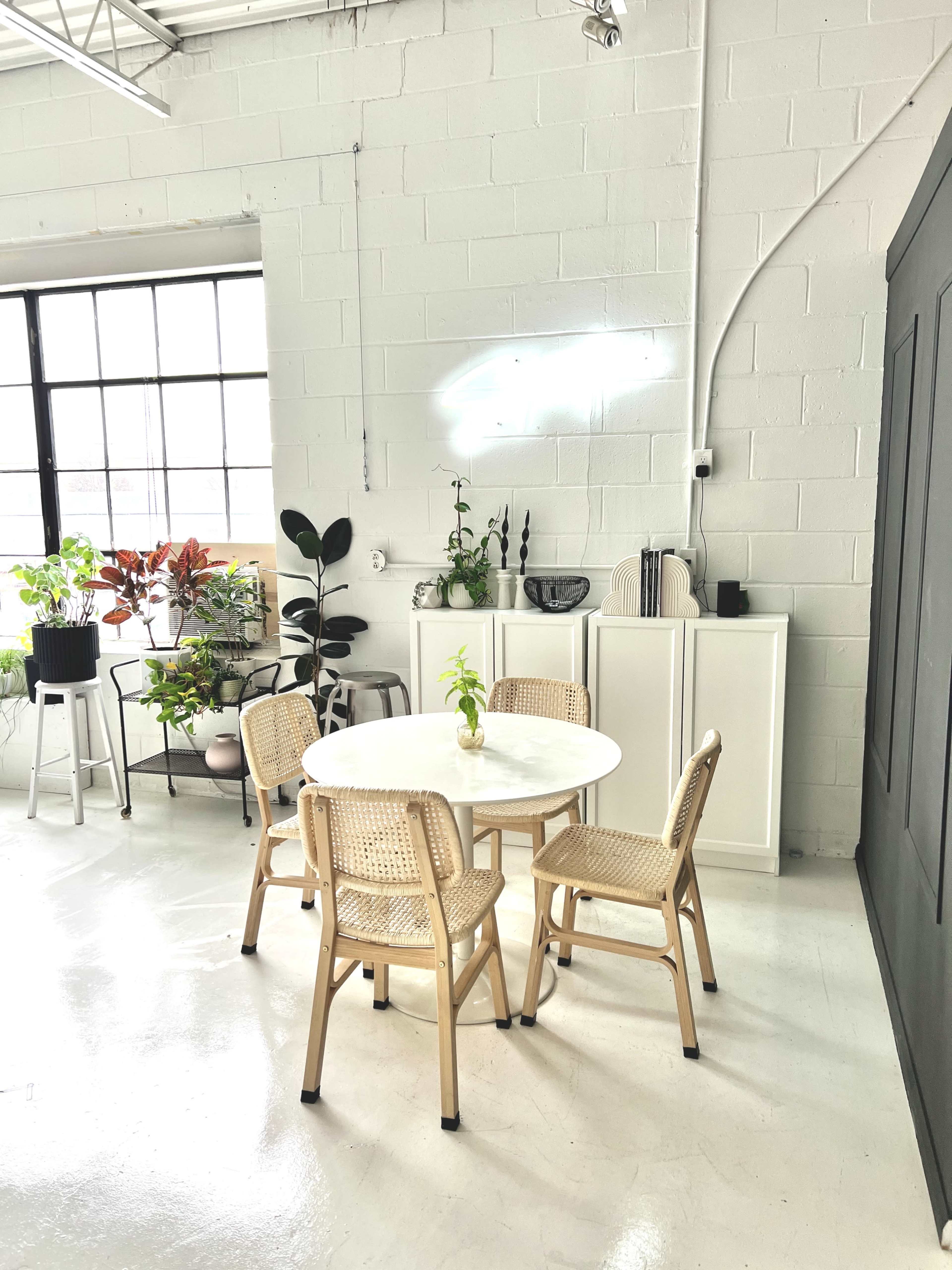 A small, round table with four rattan chairs is set up in a bright, minimalist room alongside potted plants and a white cabinet.