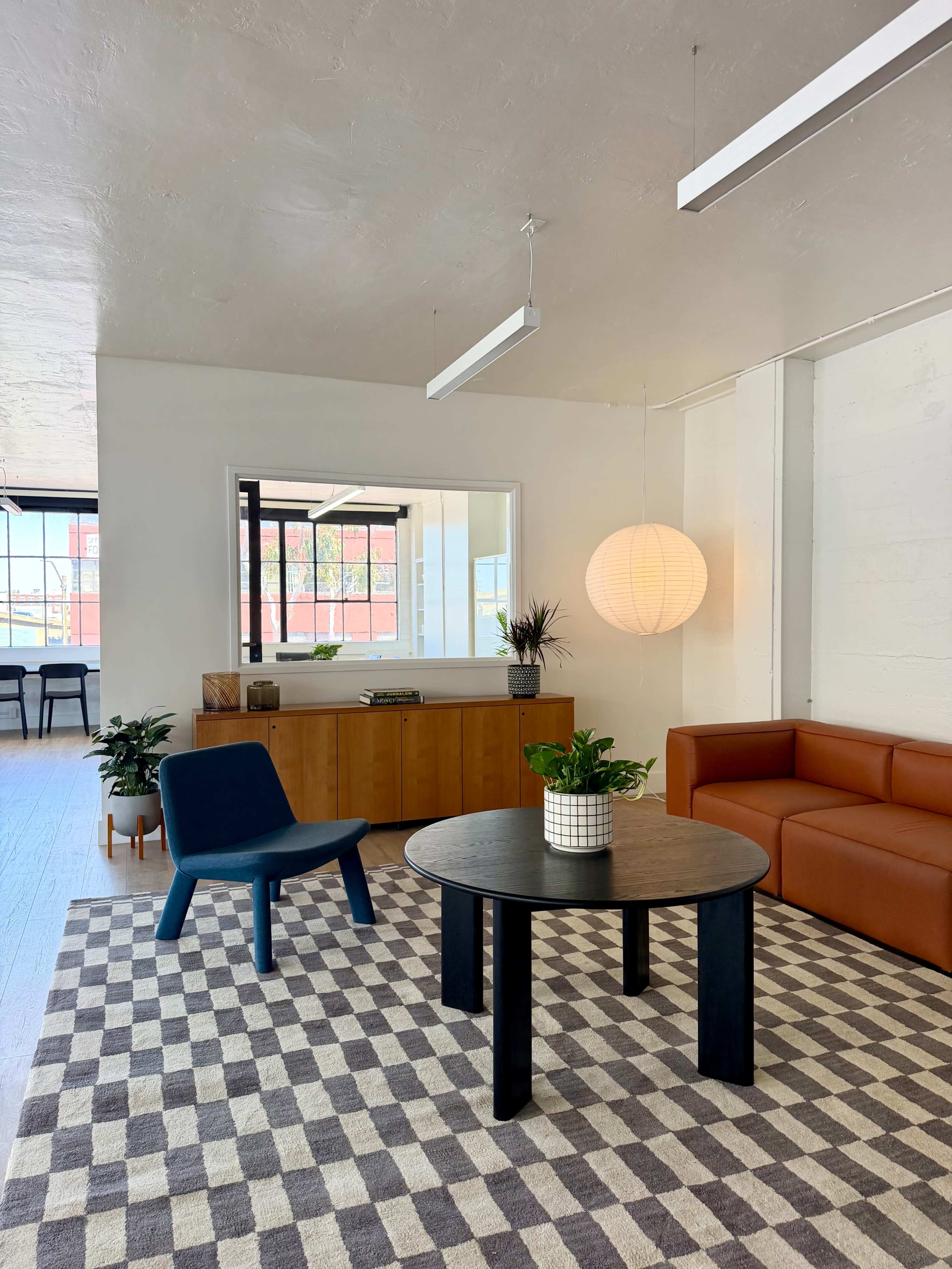 The image shows a modern, minimalist interior with a brown sectional sofa, a round black table, a blue chair, and a light fixture hanging from the ceiling.