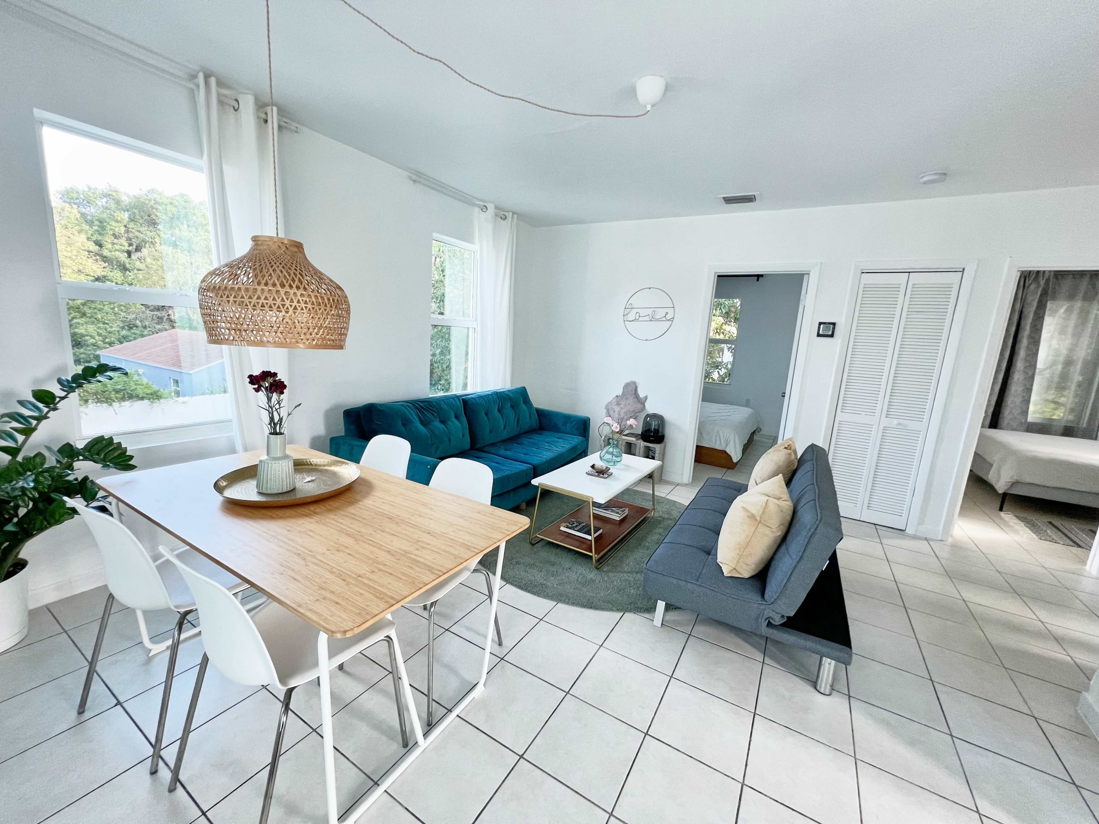 The image shows a bright, open living area featuring a dining table, two sofas in different colors, and large windows allowing natural light to enter.