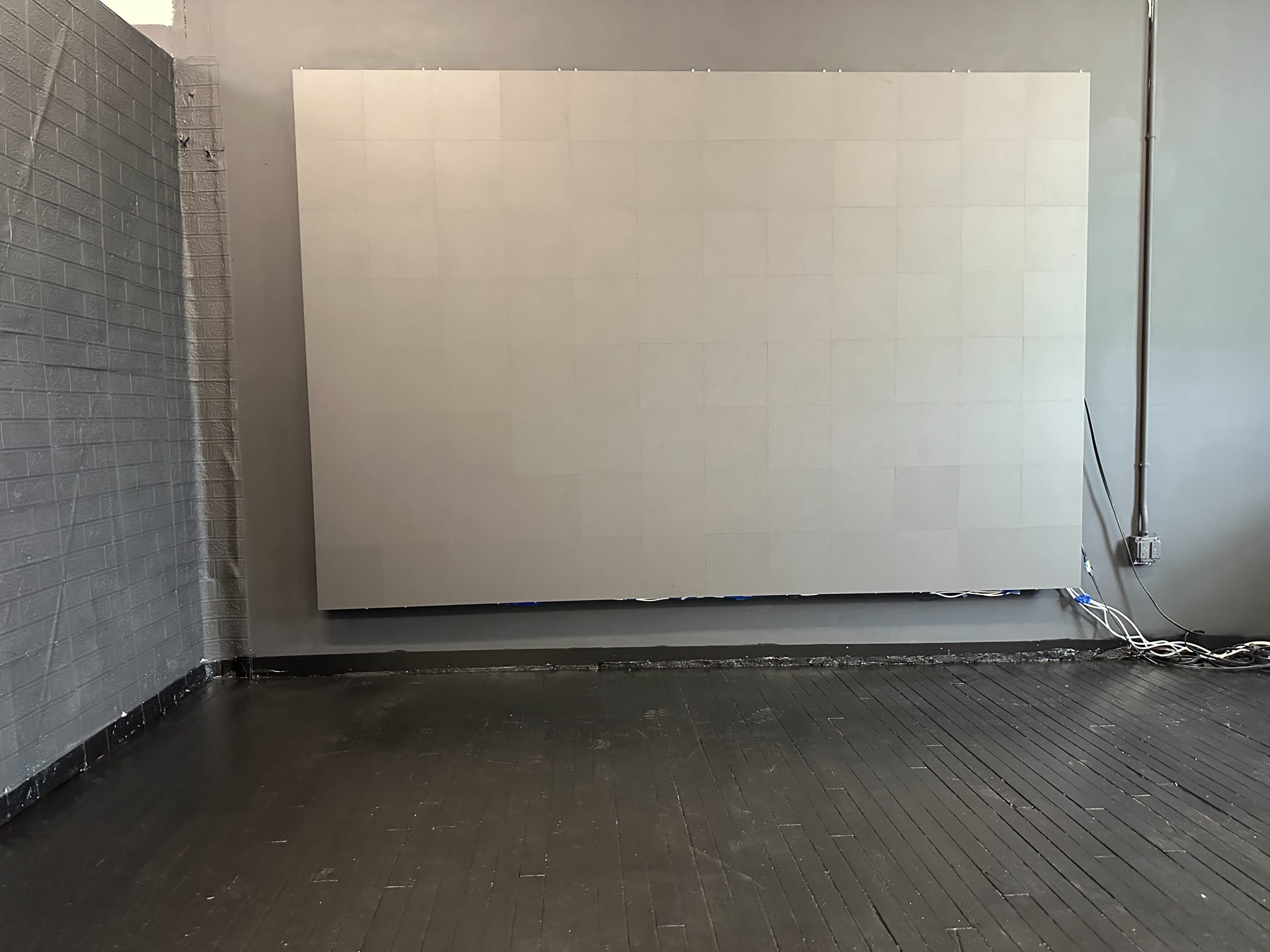 The image shows a large blank canvas mounted on a wall in a room with a dark wooden floor and exposed electrical cords.
