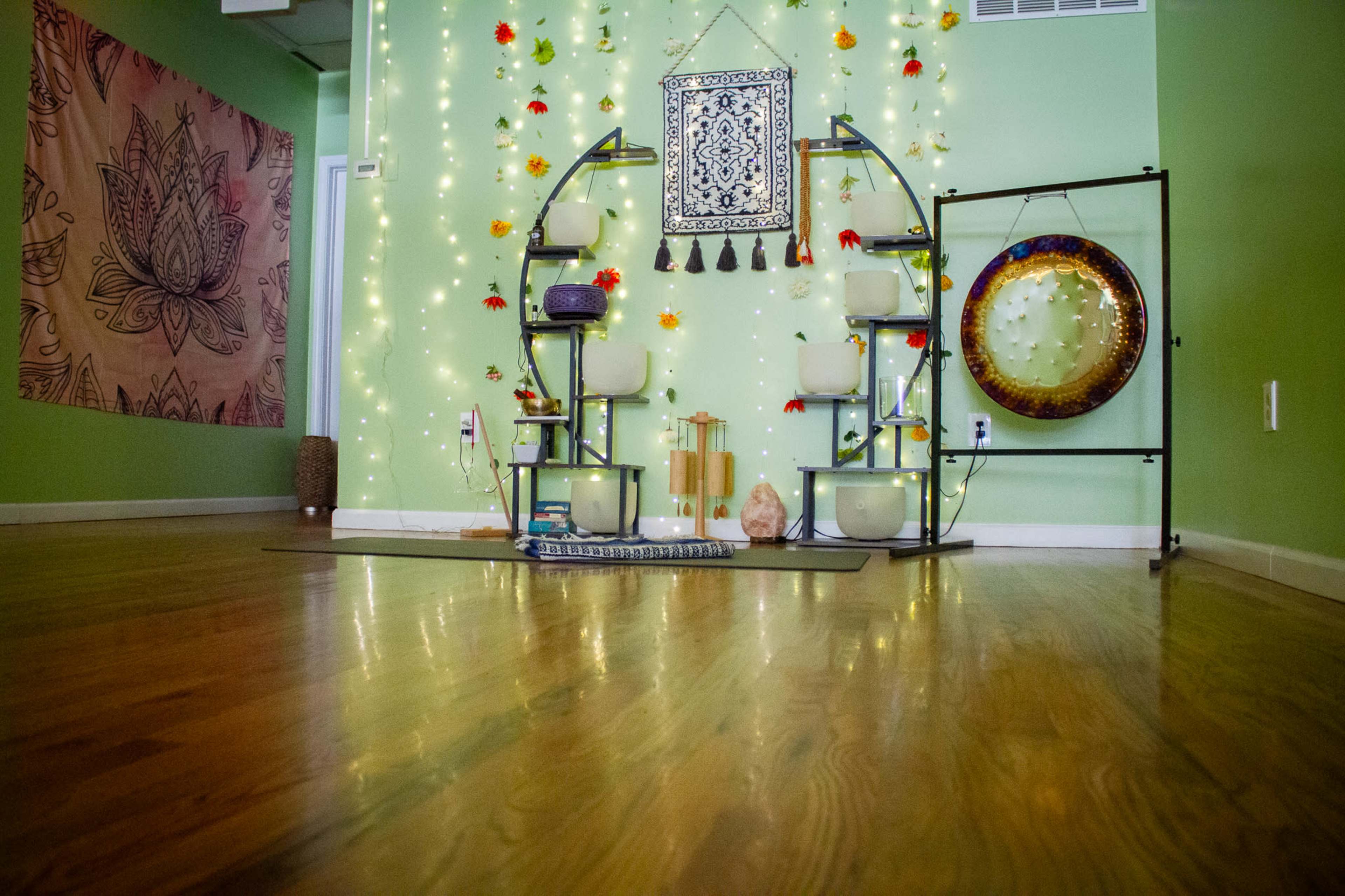 The image shows a meditation space featuring a wall adorned with decorative elements, including a patterned tapestry, floating shelves with bowls, and lighting, set on a wooden floor.