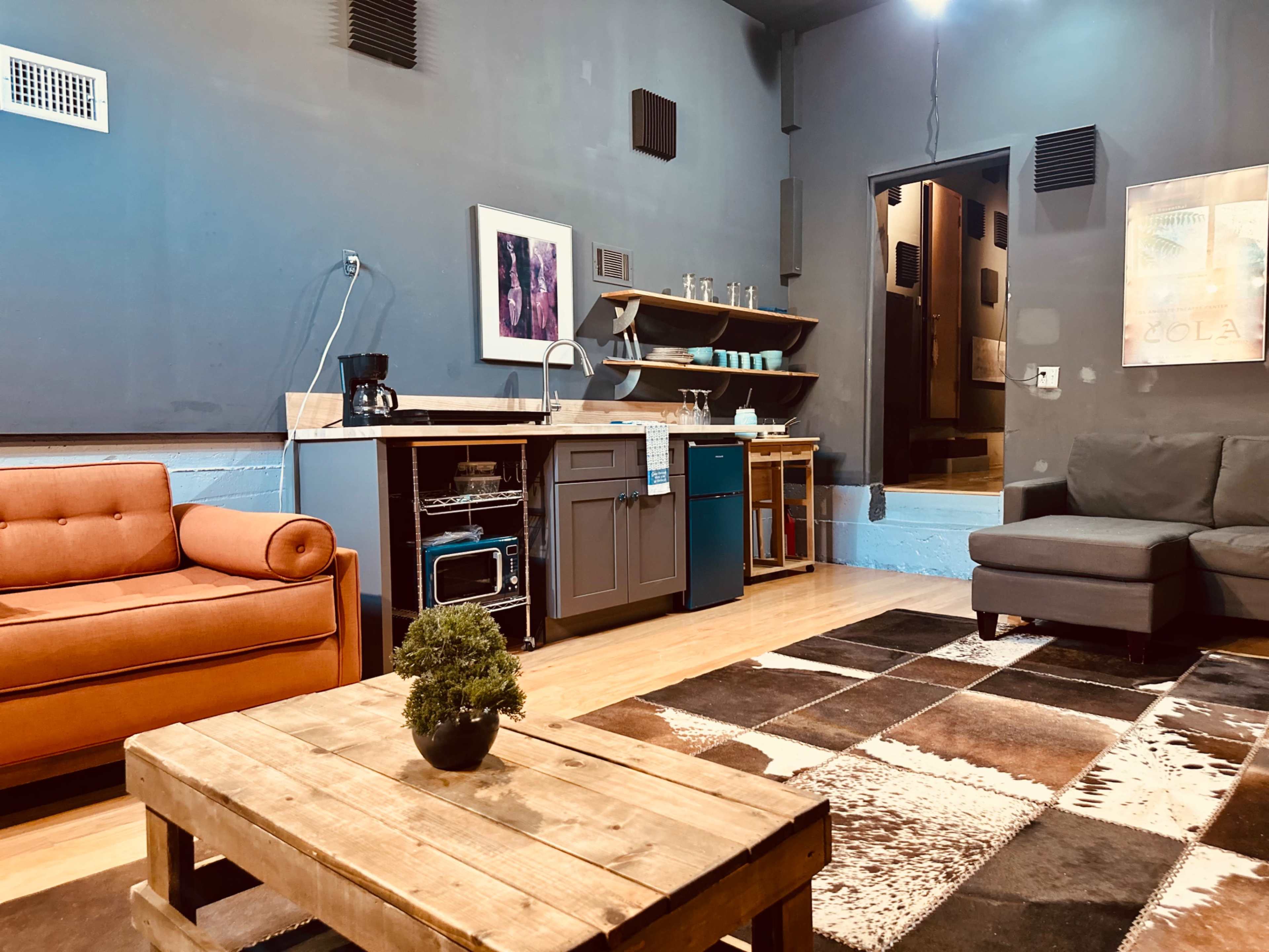The image shows a modern living space featuring a brown sofa, a dark gray couch, a wooden coffee table, and a kitchenette area with shelves and appliances.
