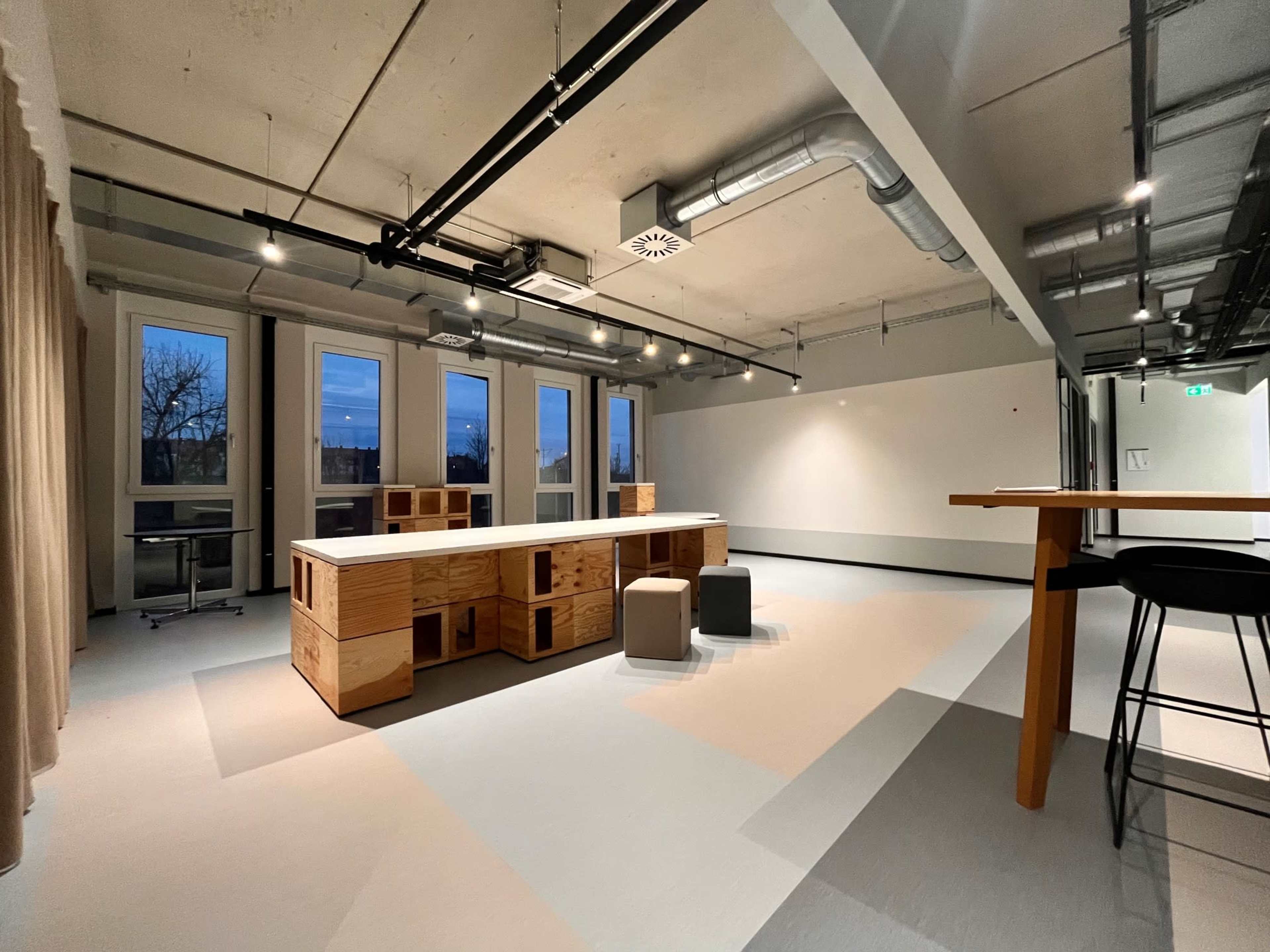 The image shows a spacious, modern interior with a central wooden island and a few stools, surrounded by large windows and minimalist decor.