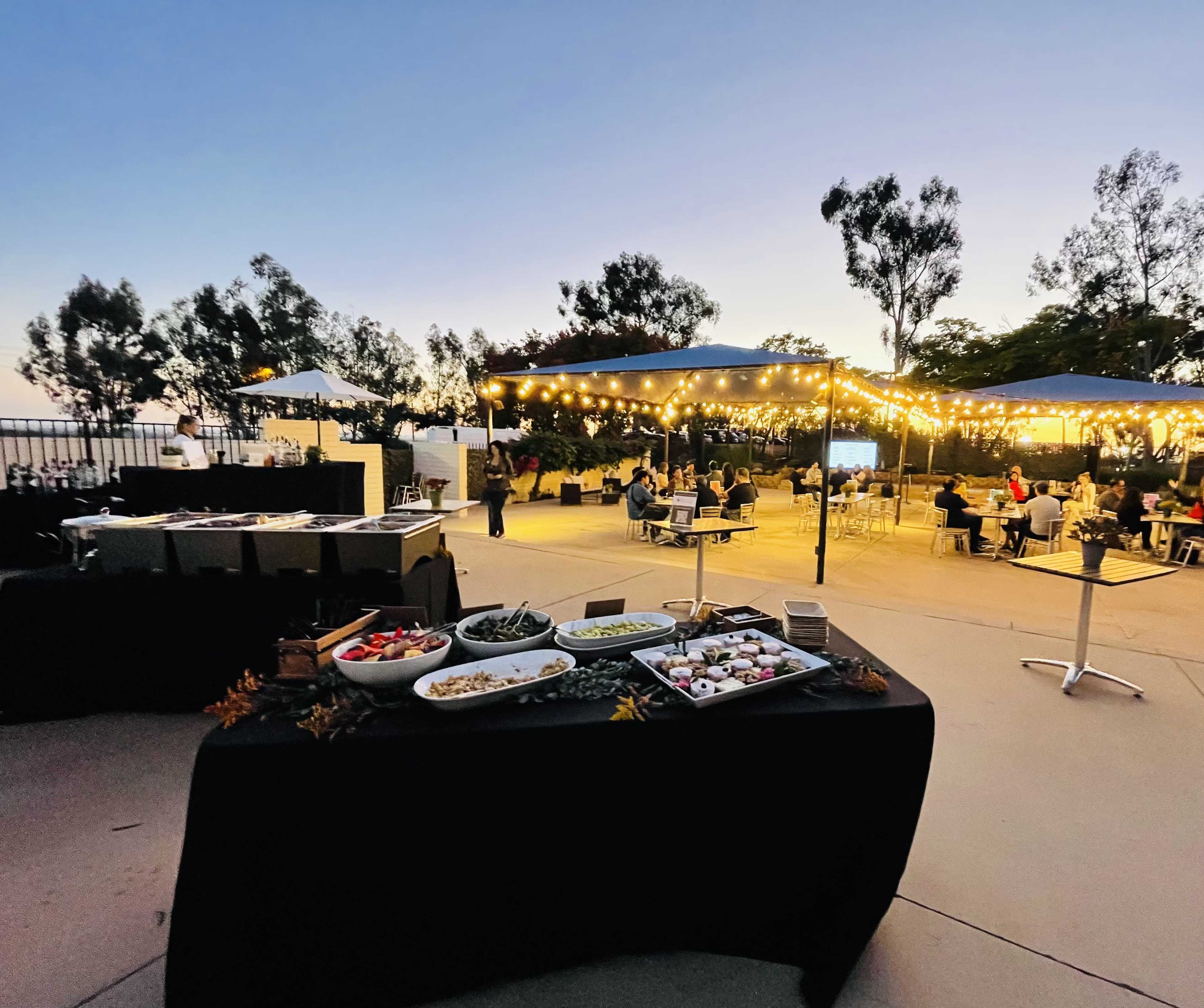 A catering table is set up with food displays under string lights as guests gather at outdoor seating areas during dusk.