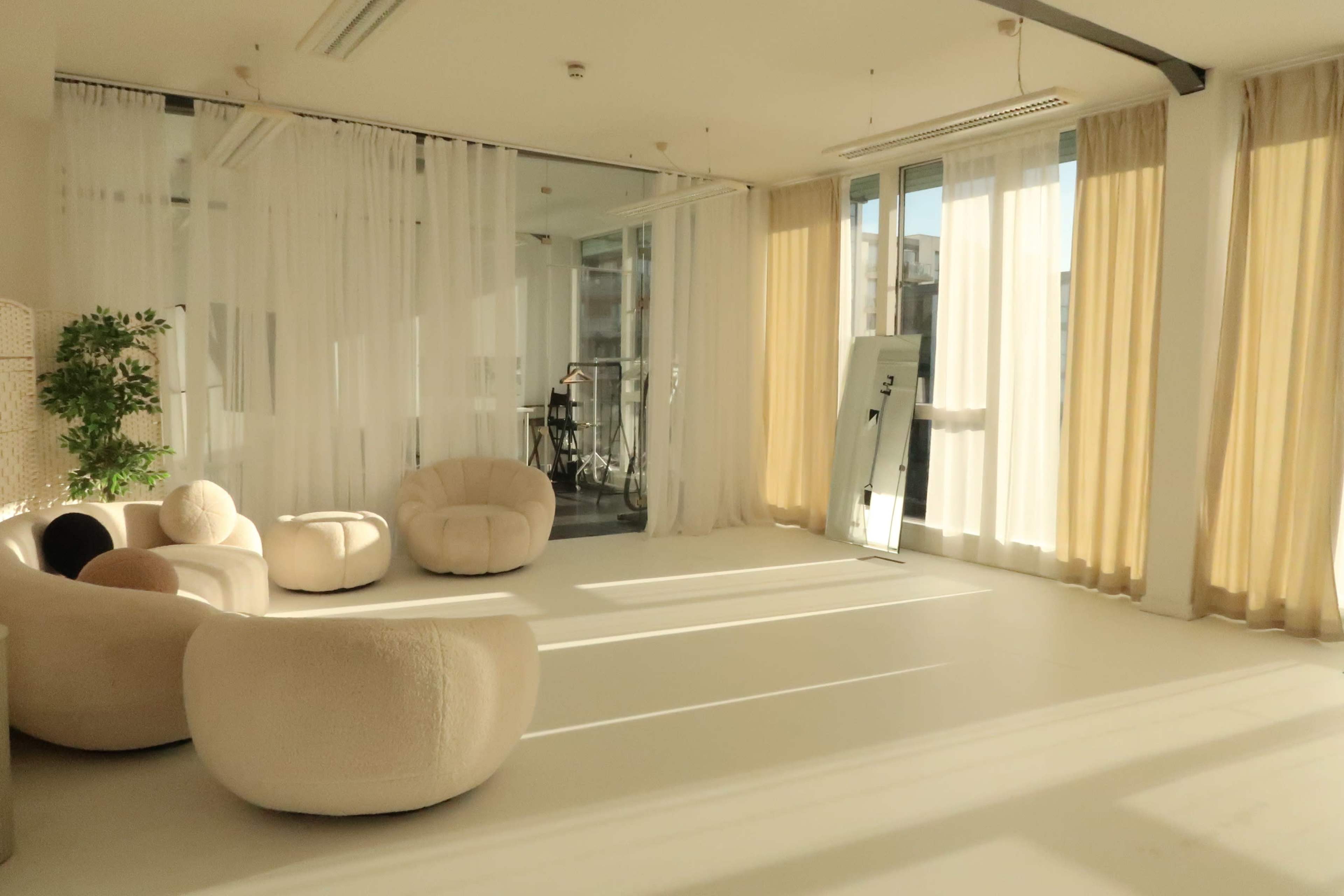 The image shows a bright, modern interior space with soft furnishings, sheer curtains, and natural light filtering through large windows.