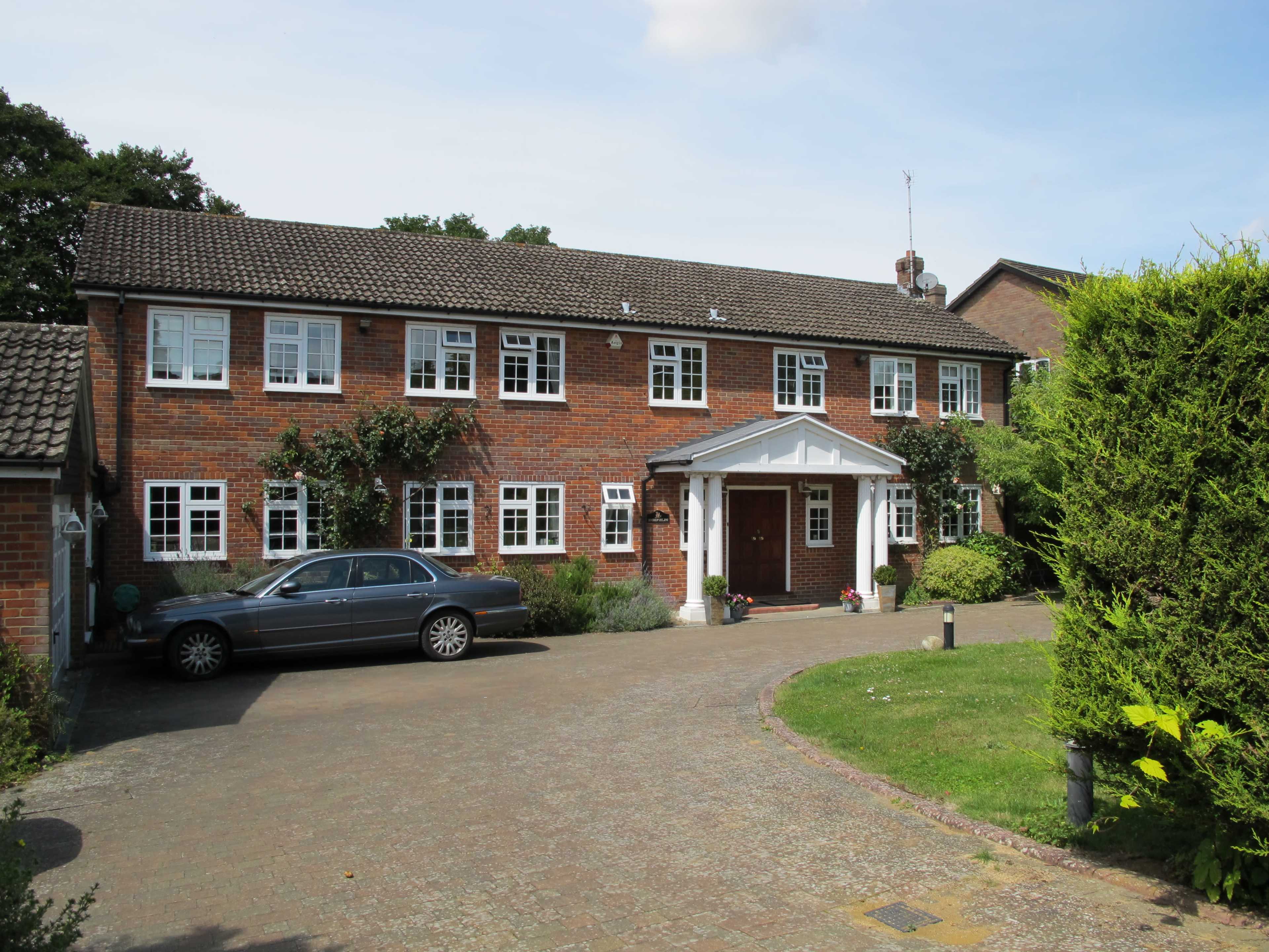 A brick house with white window frames and a covered entrance is set back from a stone driveway, alongside a parked silver car and well-kept landscaping.