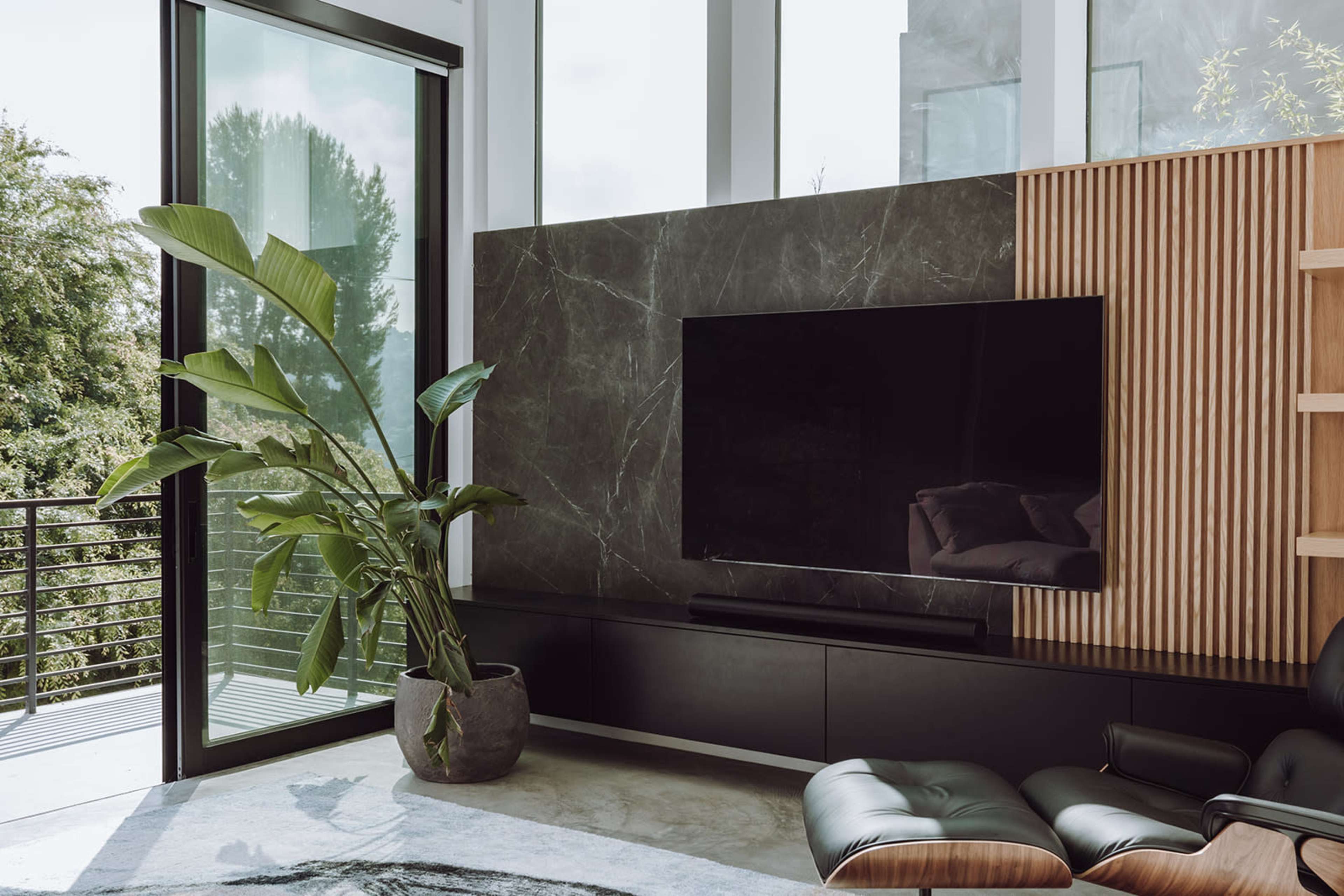 A modern living room features large windows, a dark marble accent wall with a mounted television, and a potted plant beside a sleek black media console.