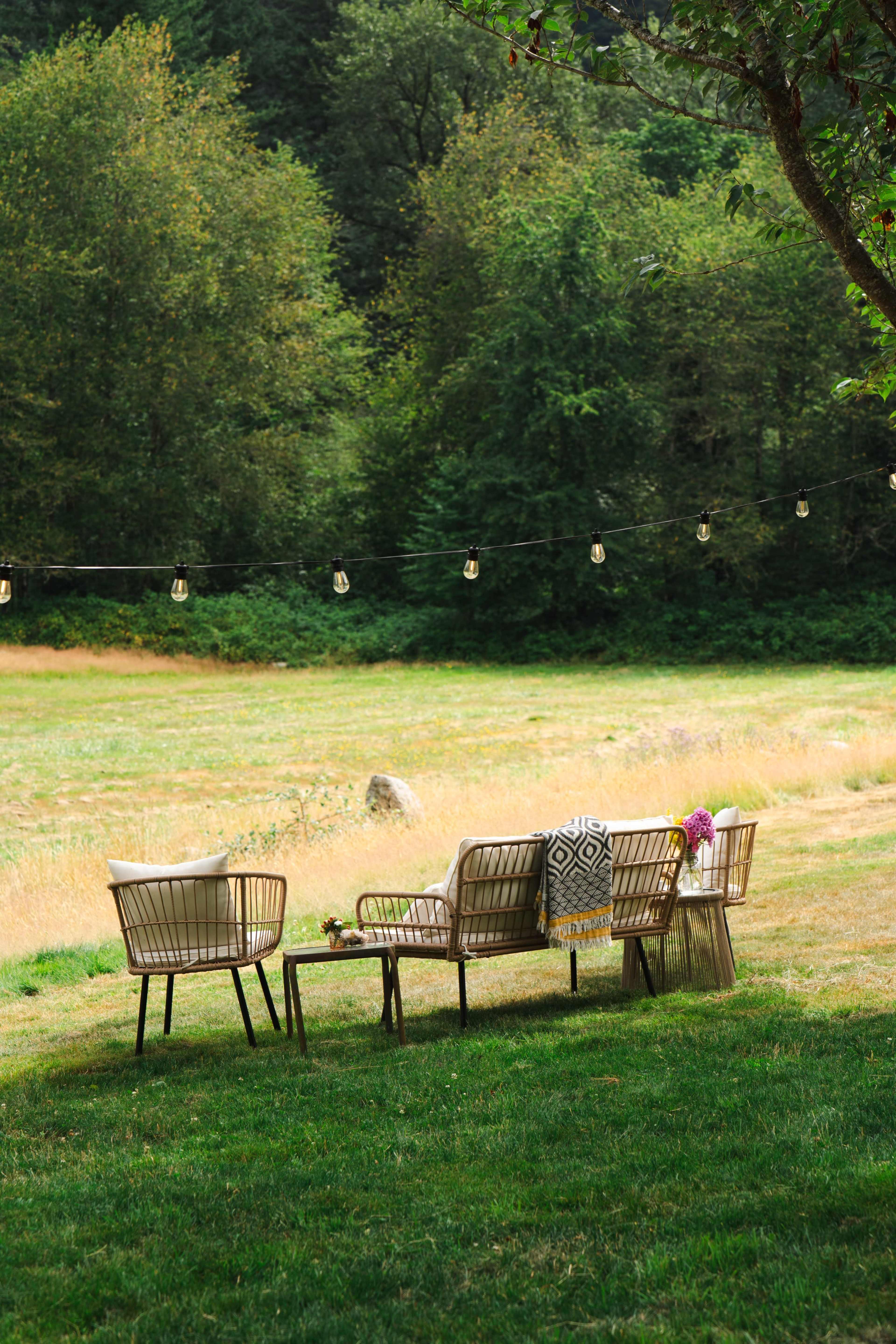 A set of outdoor wicker furniture is arranged on a grassy lawn, surrounded by trees and hanging string lights.