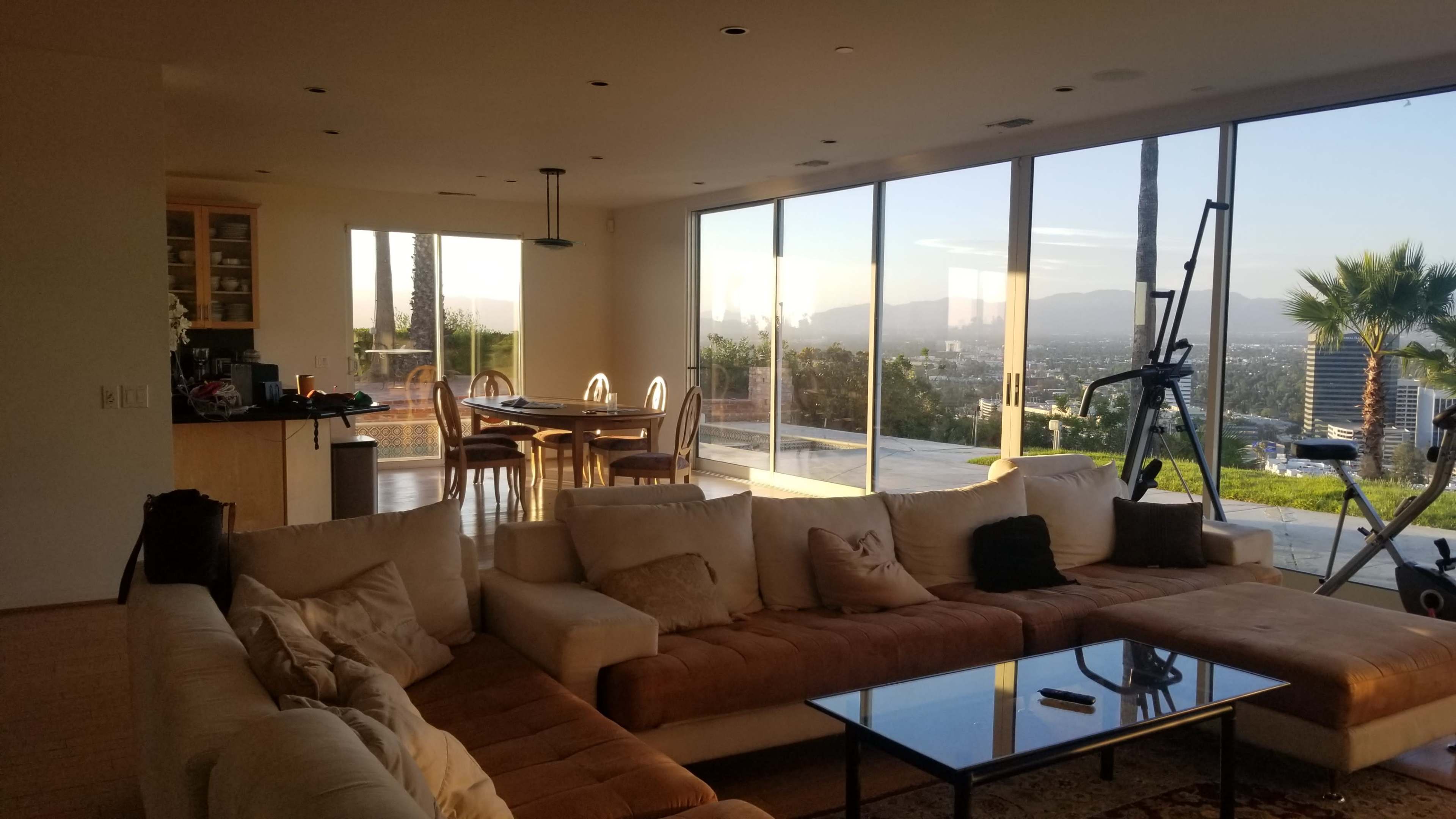 A modern living room with a large sectional sofa, a glass coffee table, and an open view of a balcony overlooking a cityscape at sunset.