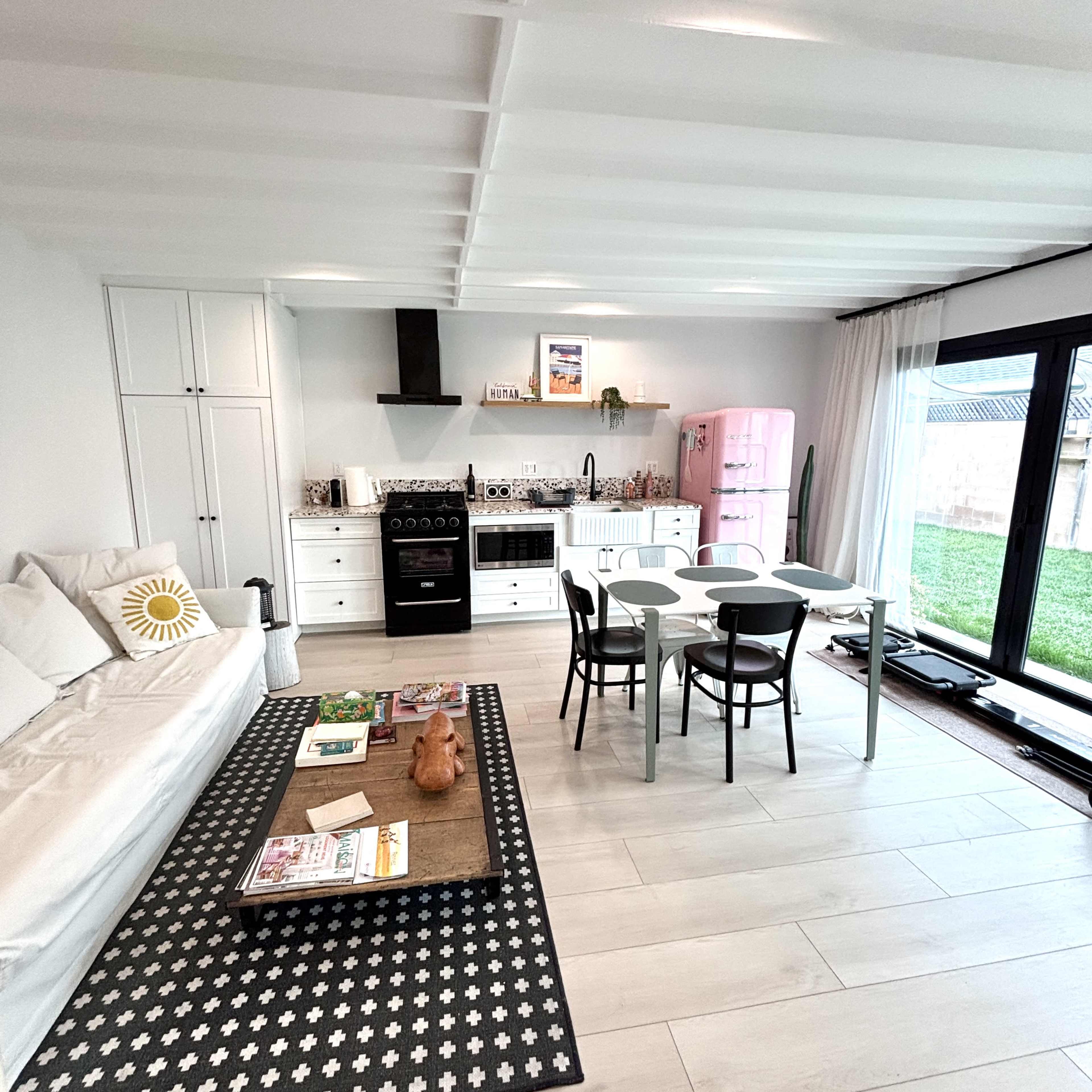 The image shows a modern kitchen and dining area with a white sofa, a black and white patterned rug, a pink refrigerator, and a dining table set for four.