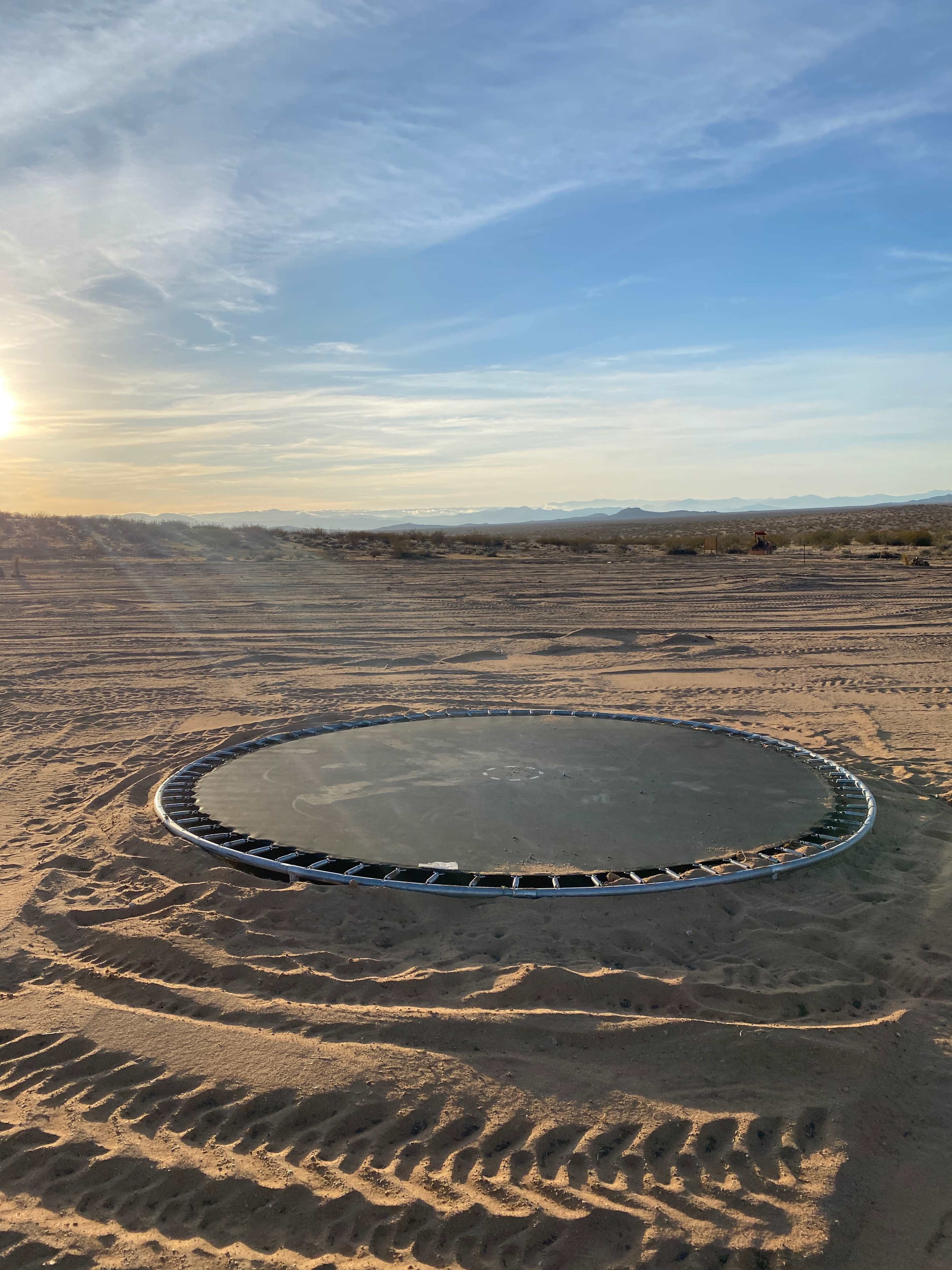 A round trampoline is positioned on a sandy desert landscape under a clear sky at sunset.