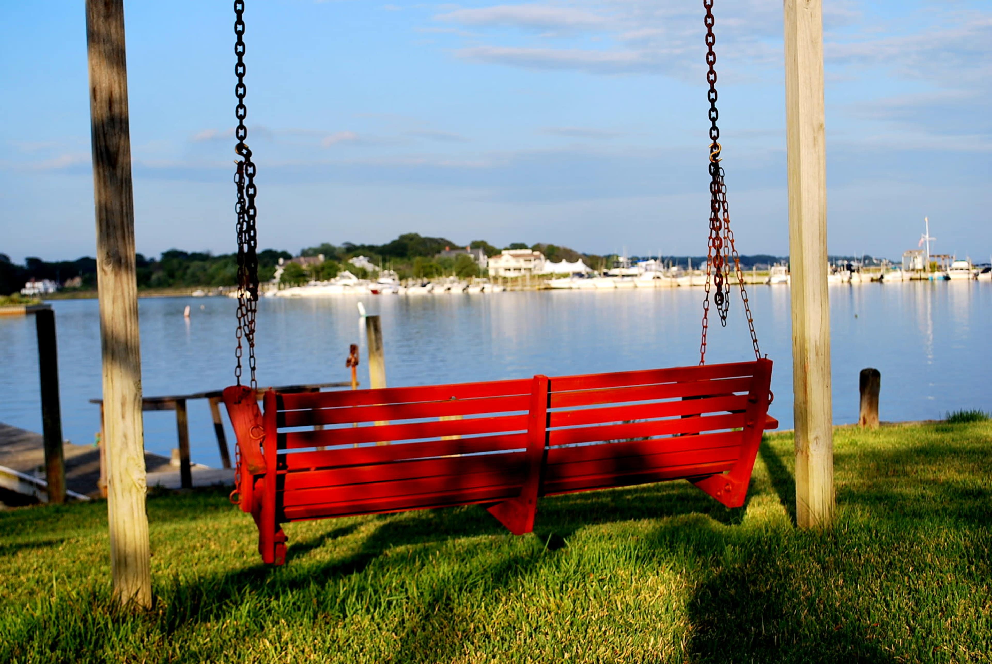 A red wooden swing hangs from a post overlooking a calm body of water with boats docked in the distance.