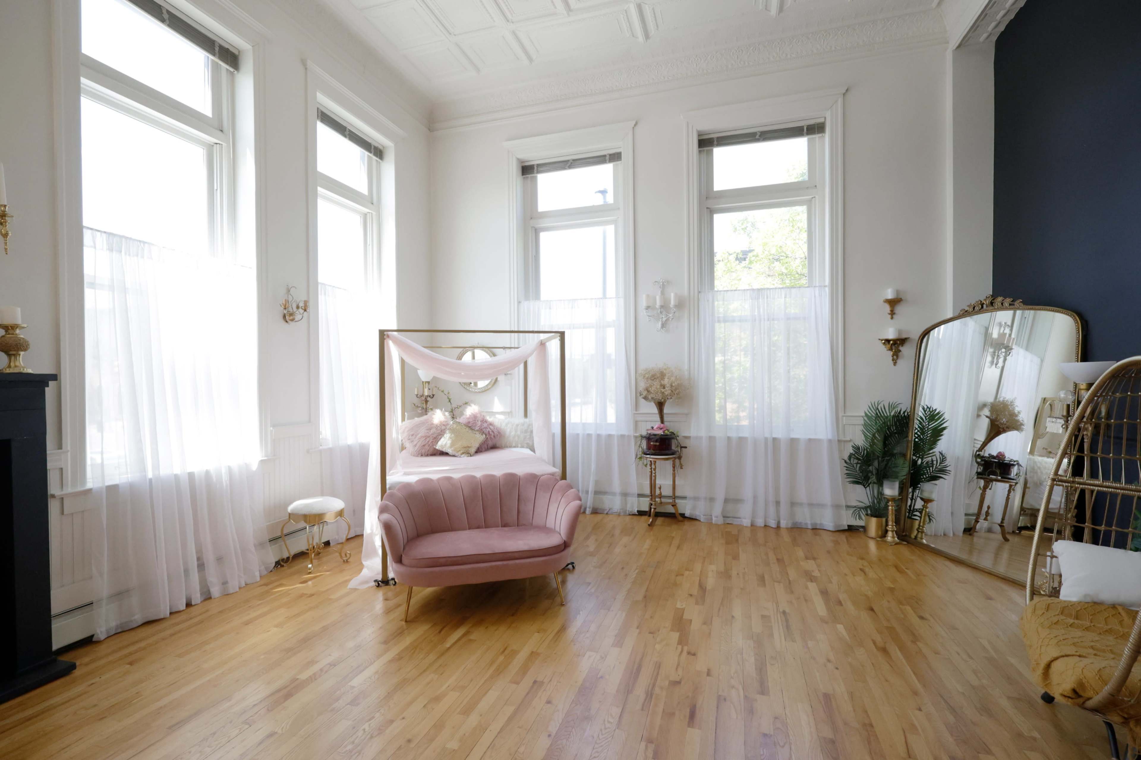 The room features a bed with a canopy, a pink velvet loveseat, large windows with sheer curtains, and decorative plants.