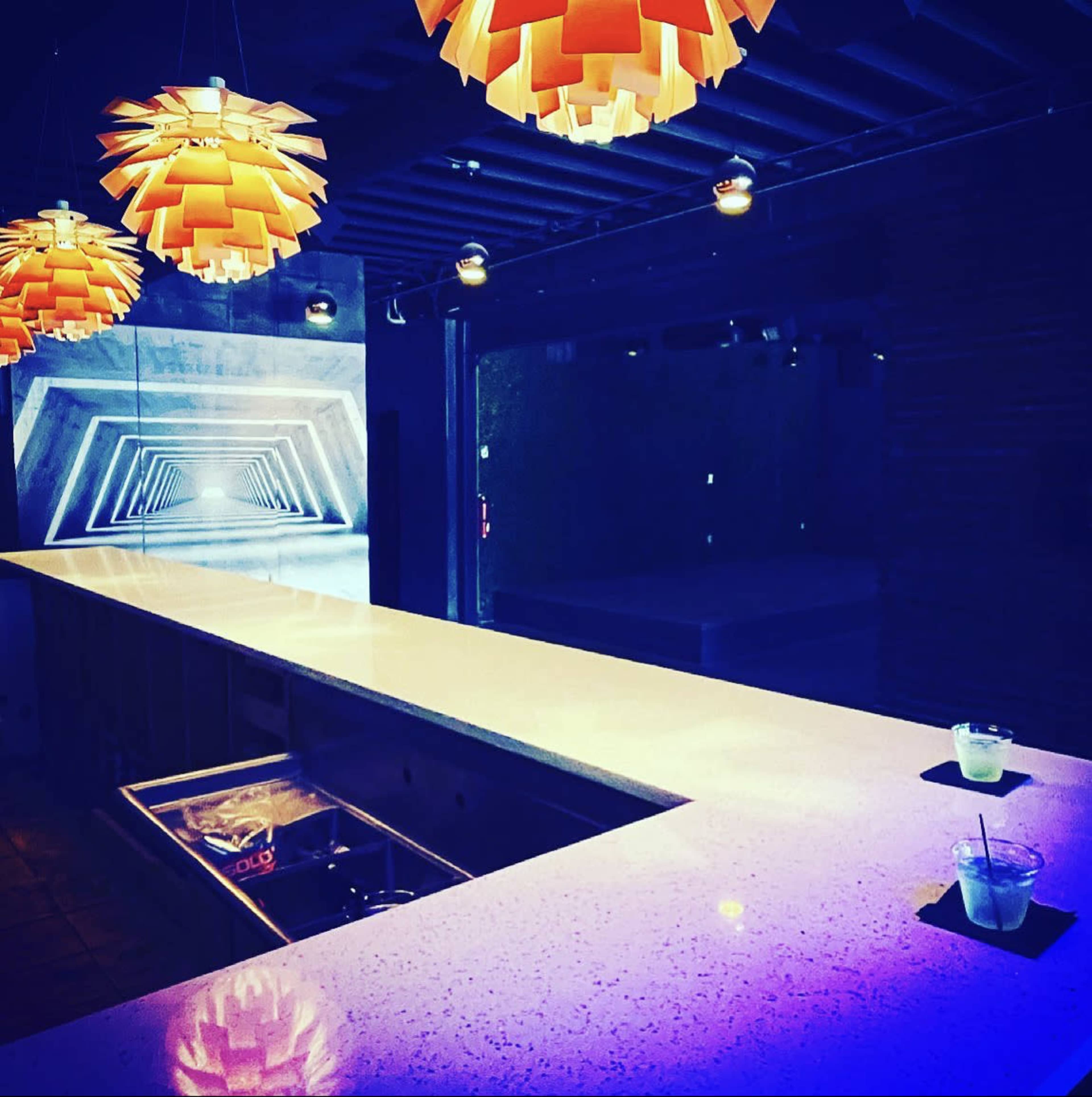 A sleek bar with a white countertop and two drinks sits under modern orange pendant lights, while illuminated geometric designs are visible on the back wall.