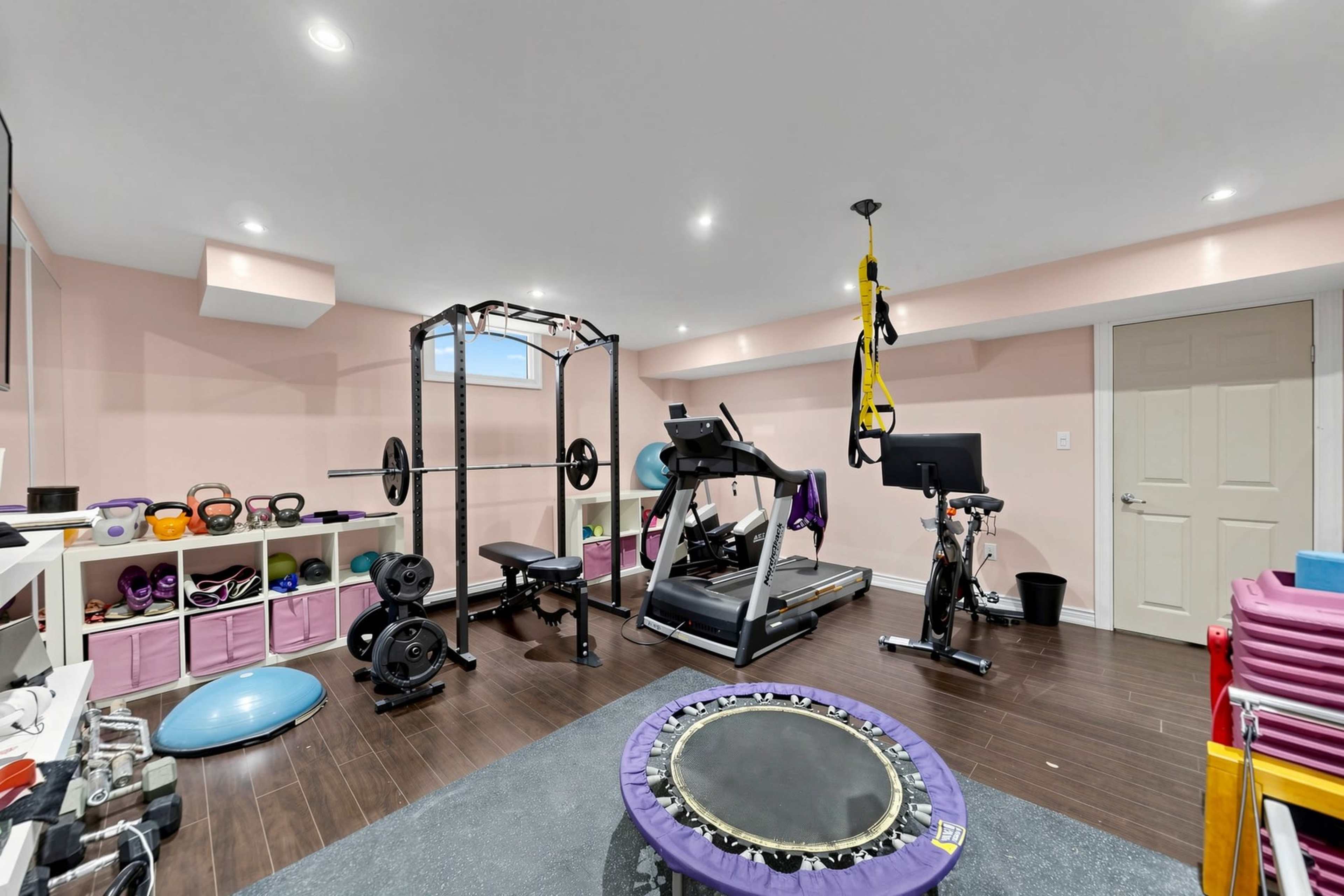 The image shows a well-equipped home gym with exercise machines, weights, and various workout accessories in a pink-walled basement space.
