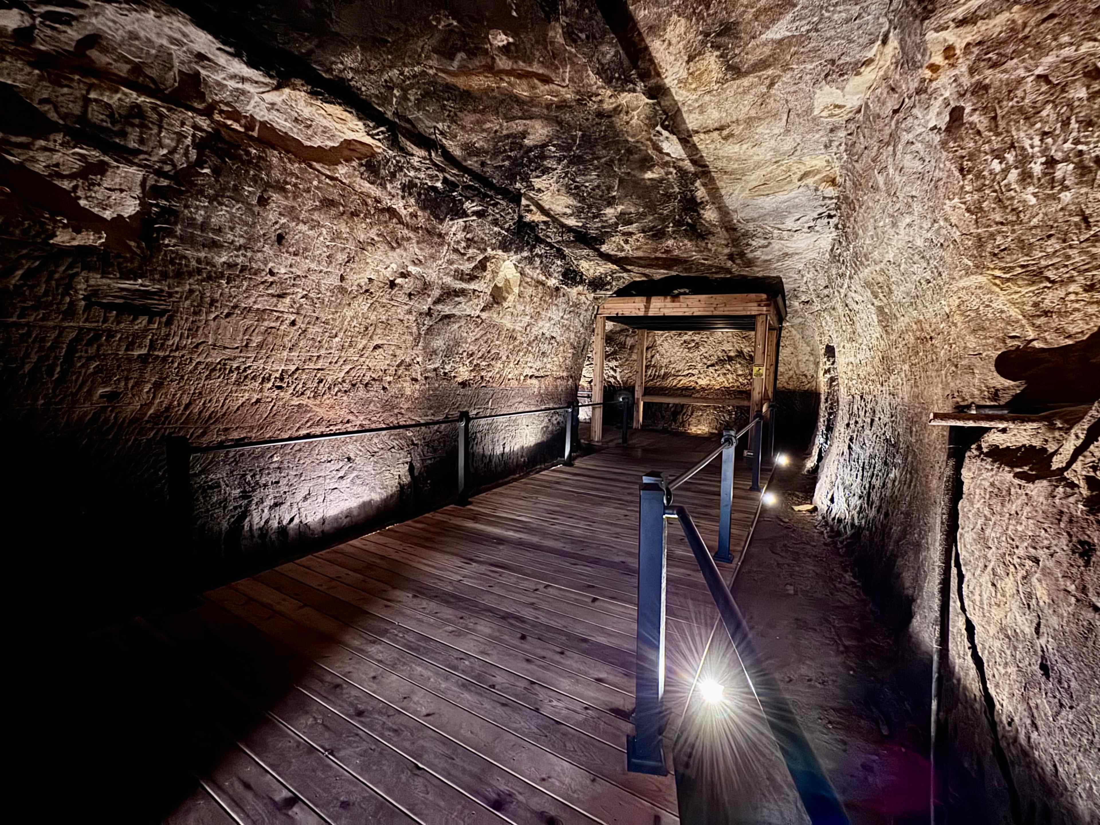 The image shows a dimly lit tunnel made of rough stone, with a wooden walkway and metal railing along the sides.